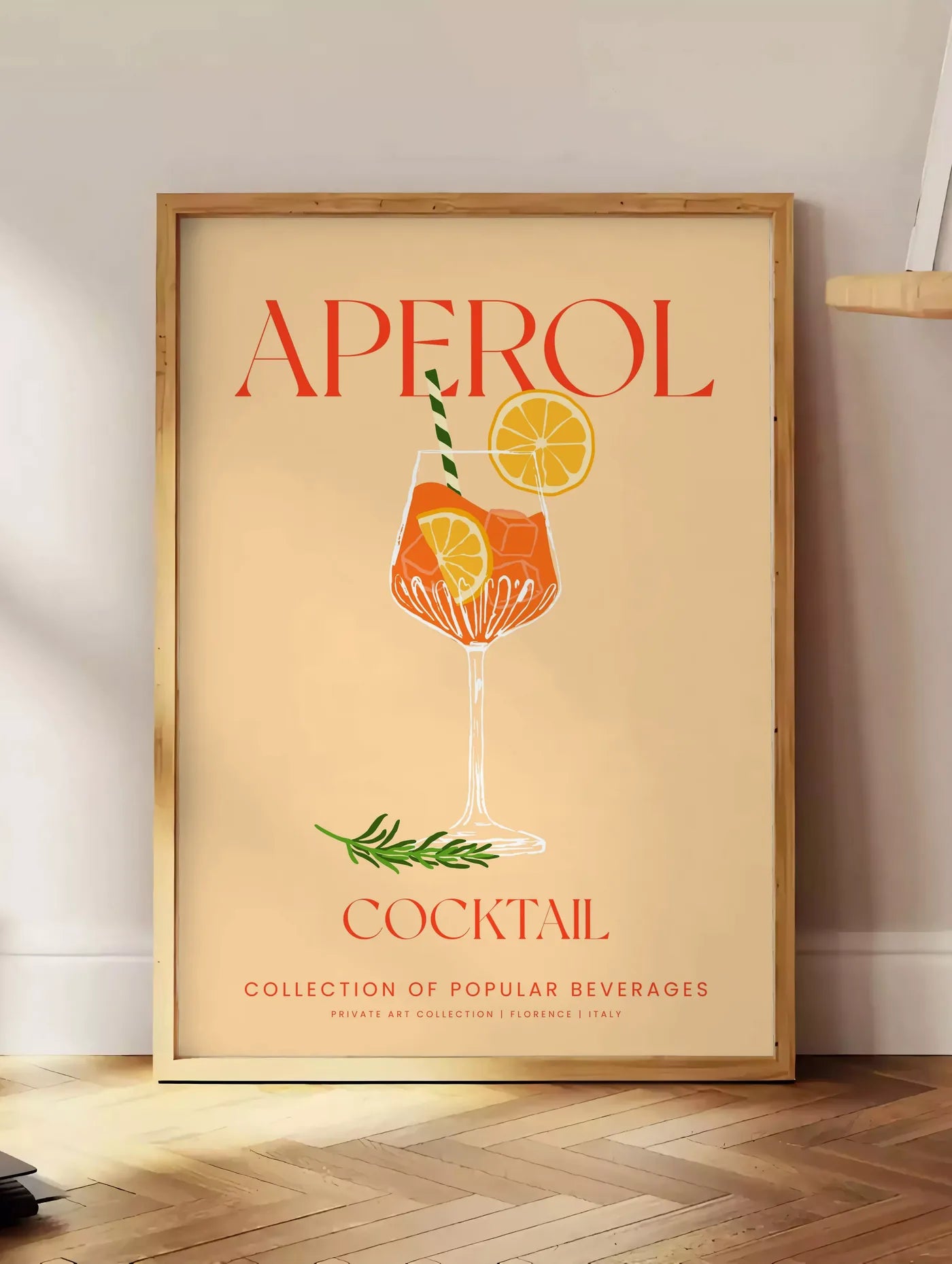 Estampe affiche cocktail Aperol Spritz, posee au sol dans un coin salon, sur un parquet a chevrons, pres d'une table d'appoint en chene chargee de livres