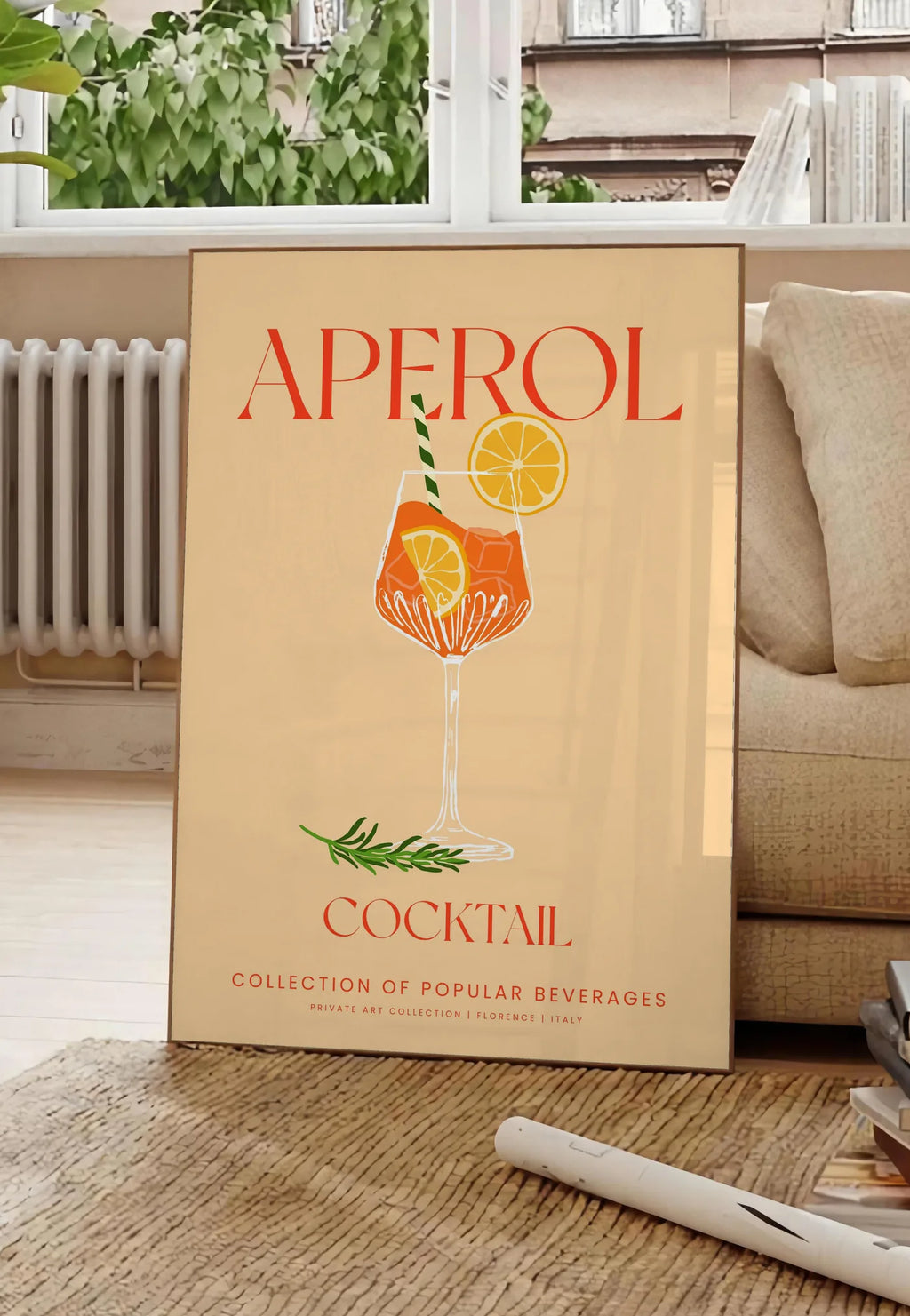 Poster affiche cocktail Aperol Spritz, posee au sol dans un salon parisien, devant une grande fenetre donnant sur un immeuble haussmannien, entre un ficus et un canape creme
