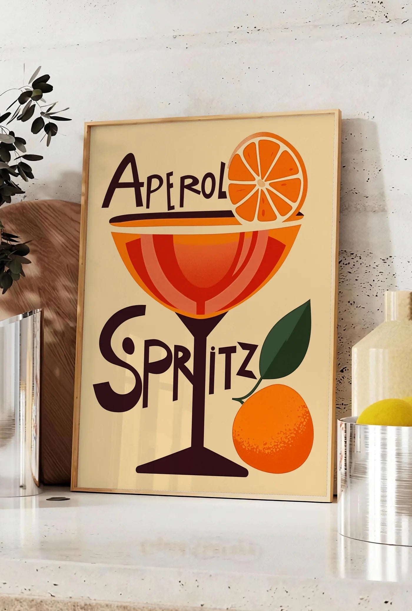 Affiche affiche cocktail Aperol Spritz, posee sur un plan de travail de cuisine en pierre claire, entre une branche d'eucalyptus en vase et un bocal de citrons