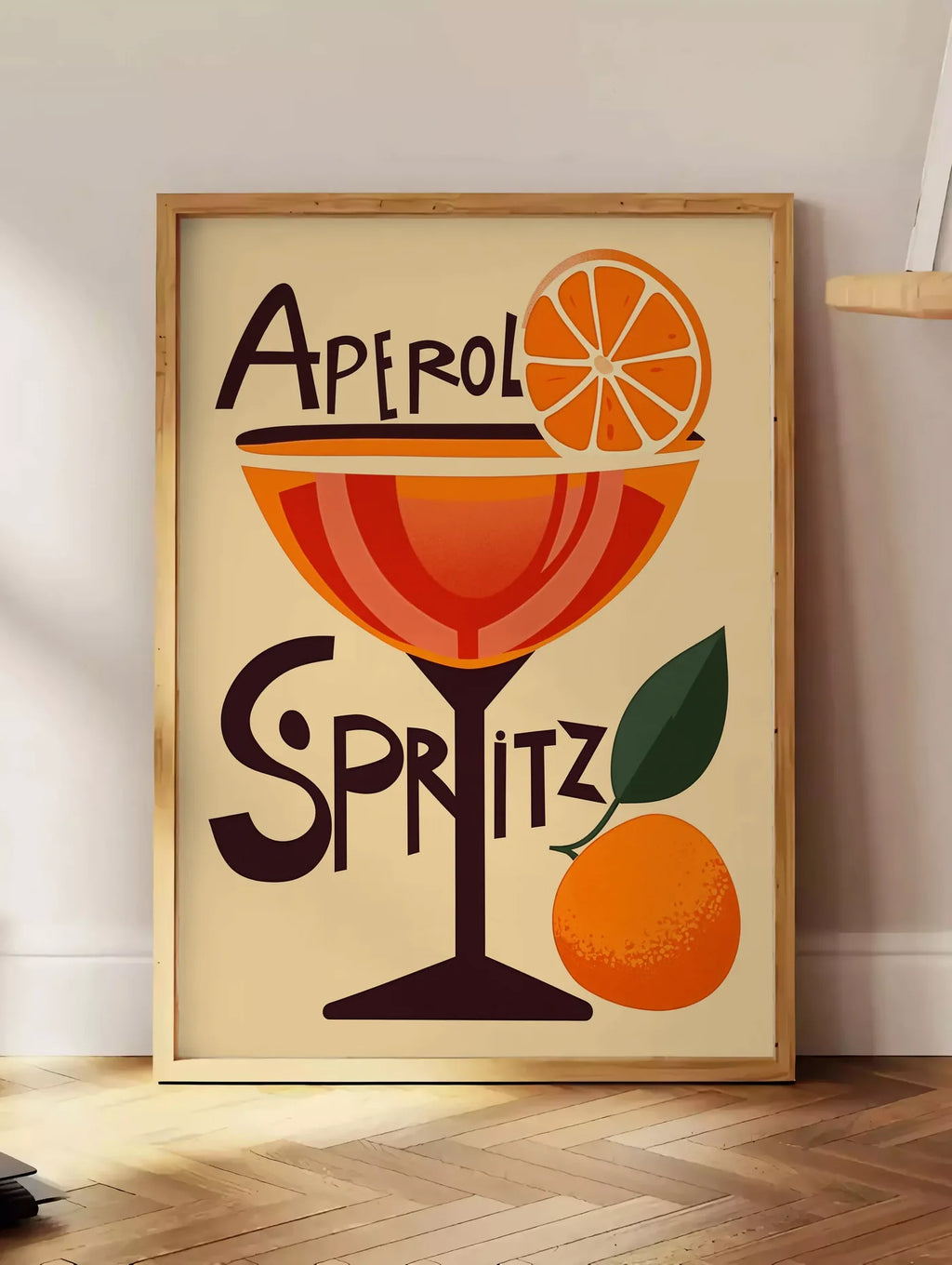 Estampe affiche cocktail Aperol Spritz, posee au sol dans un coin salon, sur un parquet a chevrons, pres d'une table d'appoint en chene chargee de livres