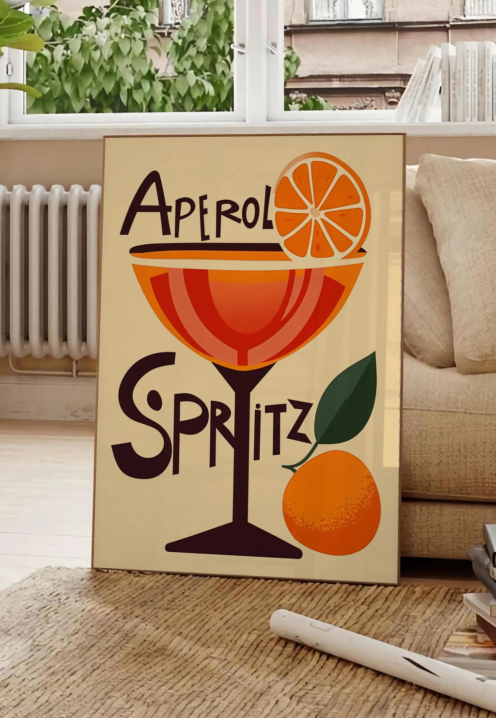 Poster affiche cocktail Aperol Spritz, posee au sol dans un salon parisien, devant une grande fenetre donnant sur un immeuble haussmannien, entre un ficus et un canape creme