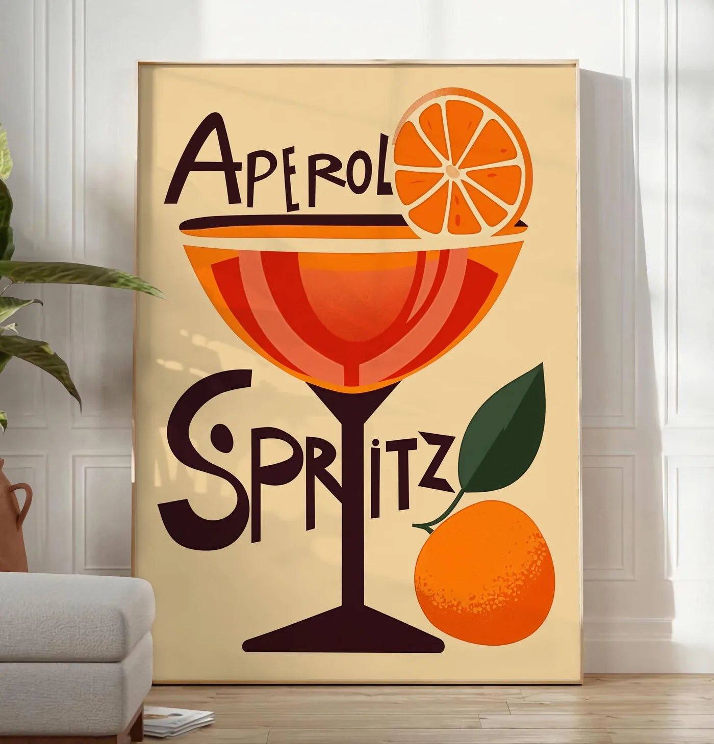 Reproduction affiche cocktail Aperol Spritz, posee au sol dans un salon minimaliste clair, a cote d'un canape beige et d'une plante verte, sur un parquet clair