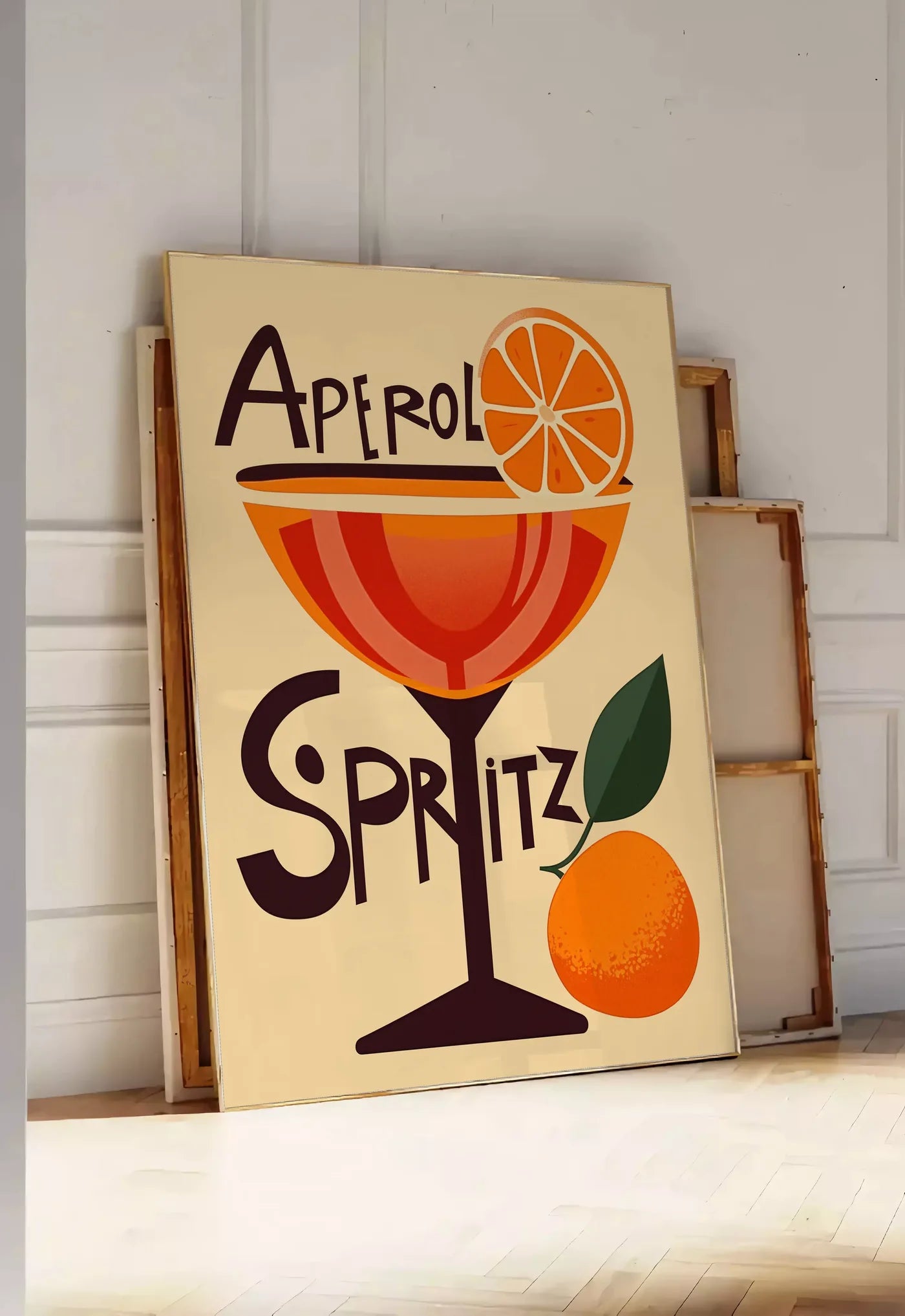 Tirage encadrable affiche cocktail Aperol Spritz, posee au sol dans une entree lumineuse, contre un mur a panneaux blancs, a cote de chassis en bois empiles