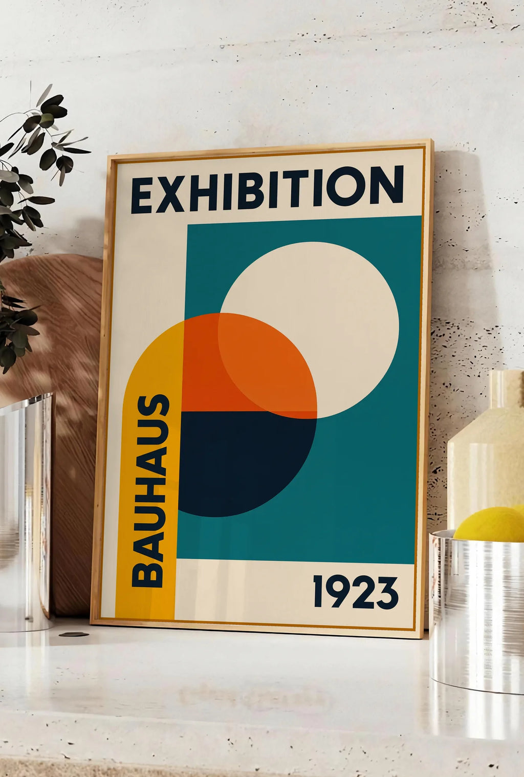 Affiche cercles superposés ocre jaune et vermillon, posee sur un plan de travail de cuisine en pierre claire, entre une branche d'eucalyptus en vase et un bocal de citrons