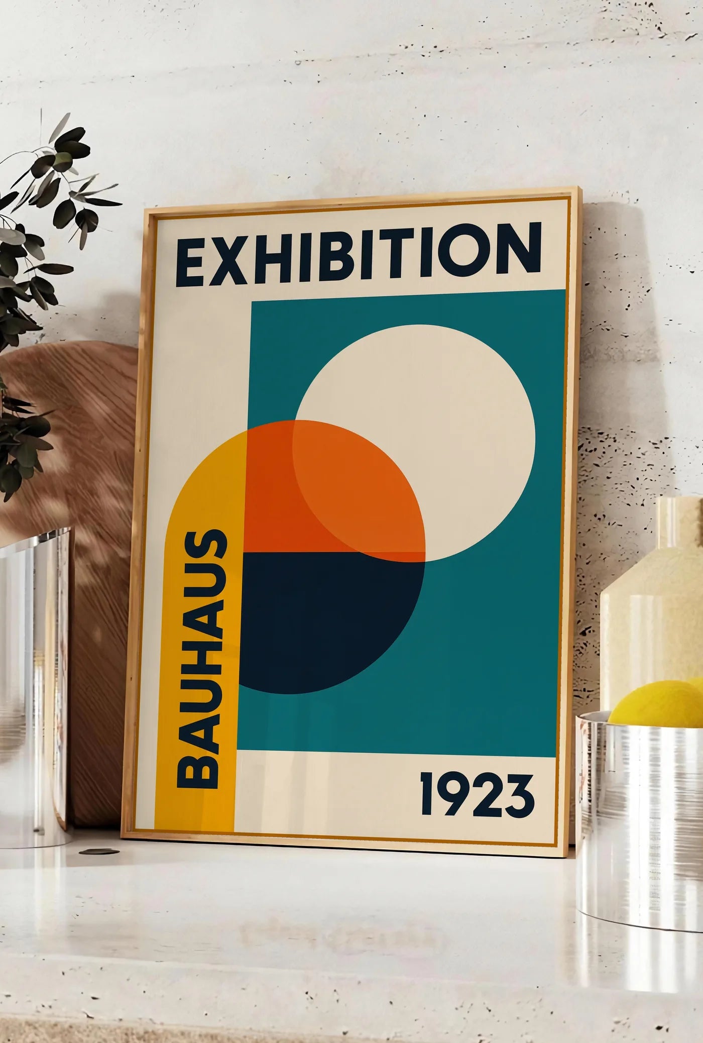 Affiche cercles superposés ocre jaune et vermillon, posee sur un plan de travail de cuisine en pierre claire, entre une branche d'eucalyptus en vase et un bocal de citrons