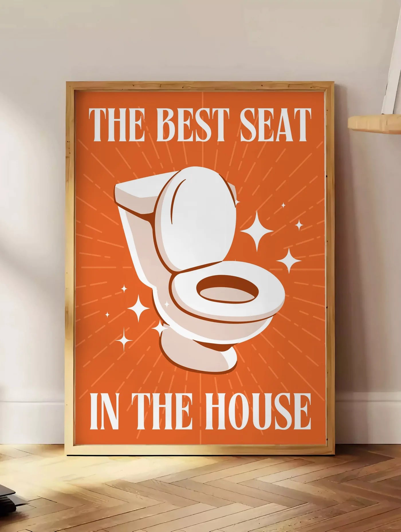 Estampe affiche toilettes humour, posee au sol dans un coin salon, sur un parquet a chevrons, pres d'une table d'appoint en chene chargee de livres