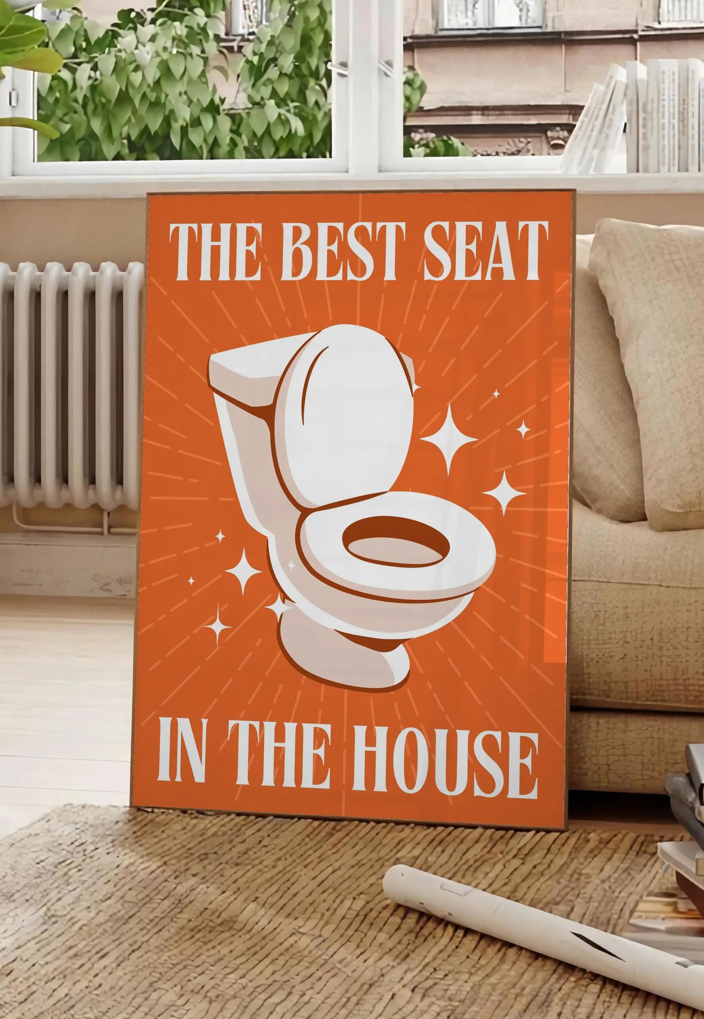 Poster affiche toilettes humour, posee au sol dans un salon parisien, devant une grande fenetre donnant sur un immeuble haussmannien, entre un ficus et un canape creme