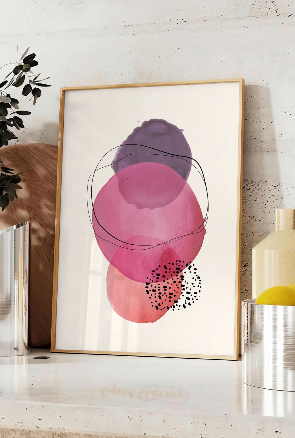 Affiche cercles aquarelle roses magenta et violets superposes sur creme, posee sur un plan de travail de cuisine en pierre claire, entre une branche d'eucalyptus en vase et un bocal de citrons