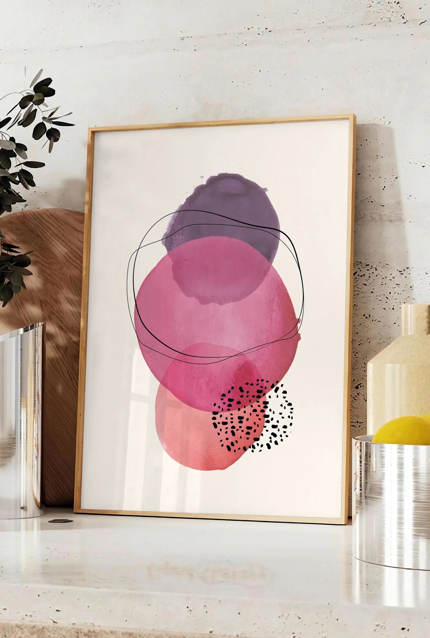 Affiche cercles aquarelle roses magenta et violets superposes sur creme, posee sur un plan de travail de cuisine en pierre claire, entre une branche d'eucalyptus en vase et un bocal de citrons