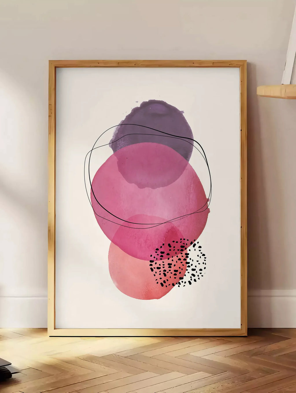 Estampe cercles aquarelle roses magenta et violets superposes sur creme, posee au sol dans un coin salon, sur un parquet a chevrons, pres d'une table d'appoint en chene chargee de livres