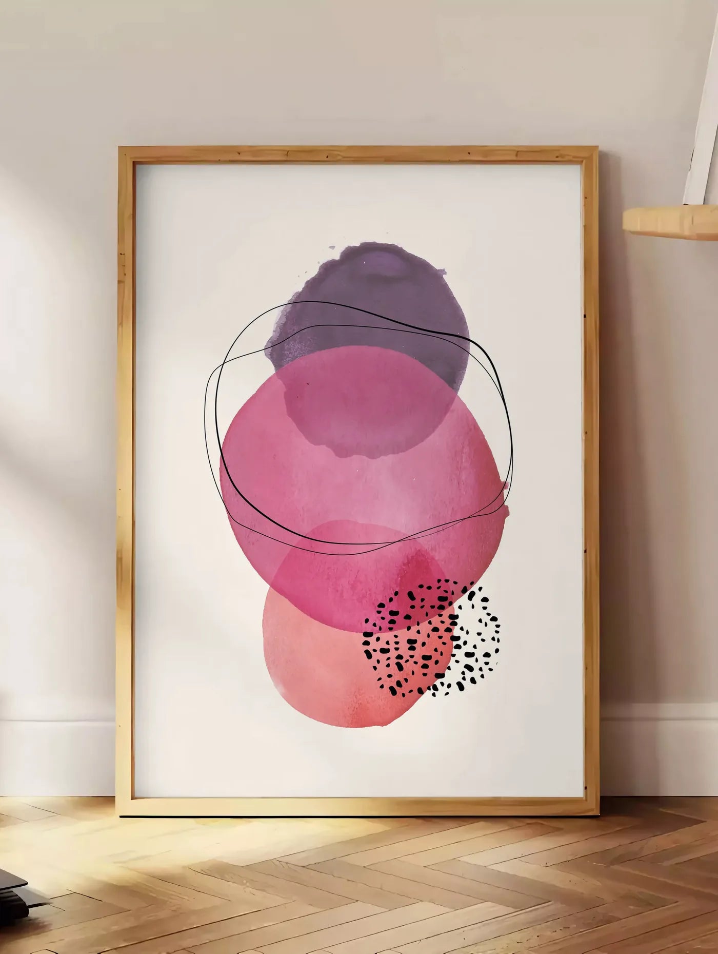 Estampe cercles aquarelle roses magenta et violets superposes sur creme, posee au sol dans un coin salon, sur un parquet a chevrons, pres d'une table d'appoint en chene chargee de livres