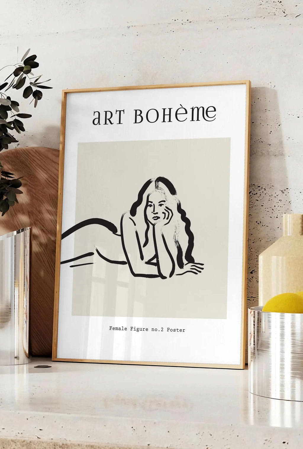 Affiche femme allongee au trait de pinceau noir sur creme, posee sur un plan de travail de cuisine en pierre claire, entre une branche d'eucalyptus en vase et un bocal de citrons