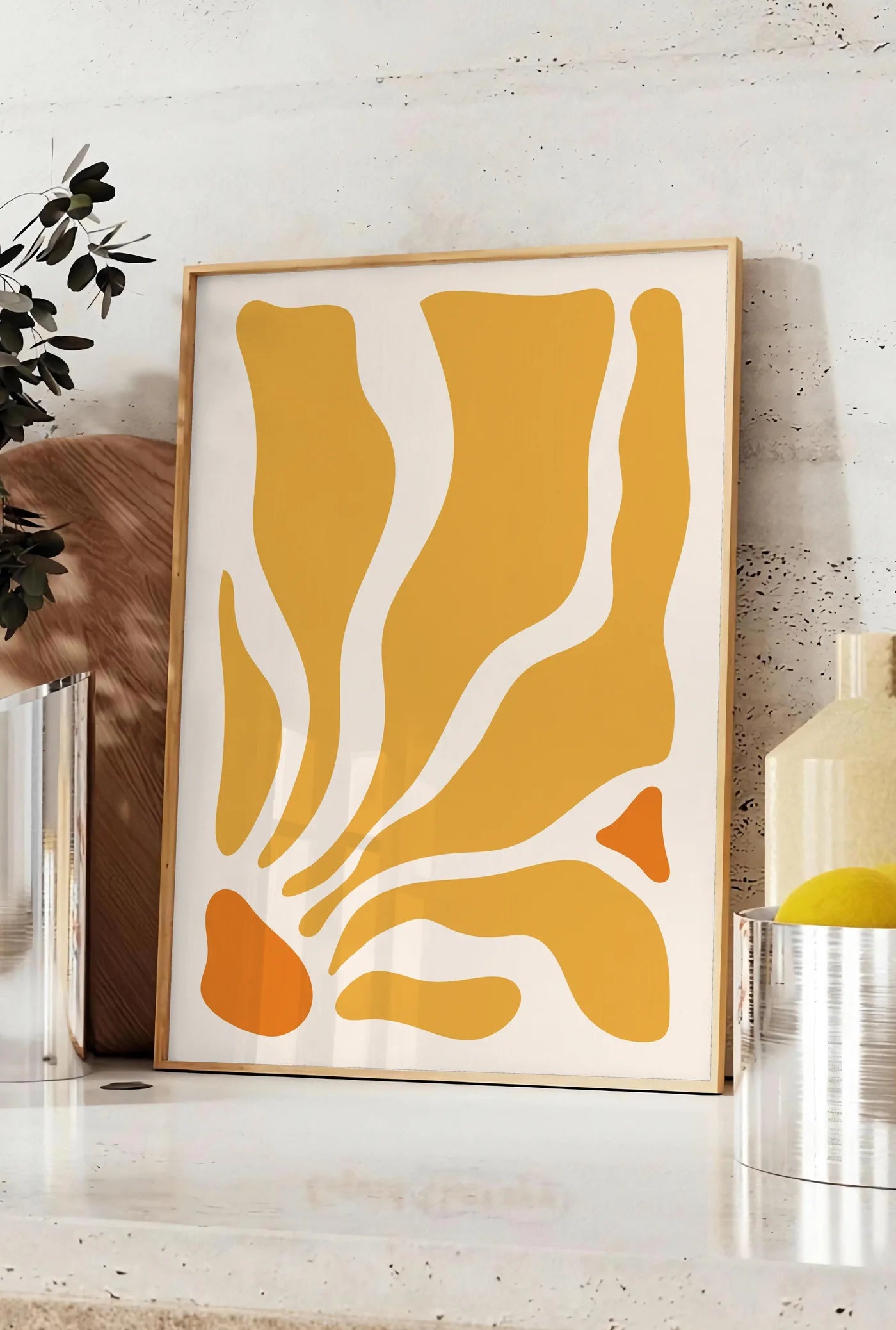Affiche formes organiques jaunes et orange ondulees sur fond creme, posee sur un plan de travail de cuisine en pierre claire, entre une branche d'eucalyptus en vase et un bocal de citrons