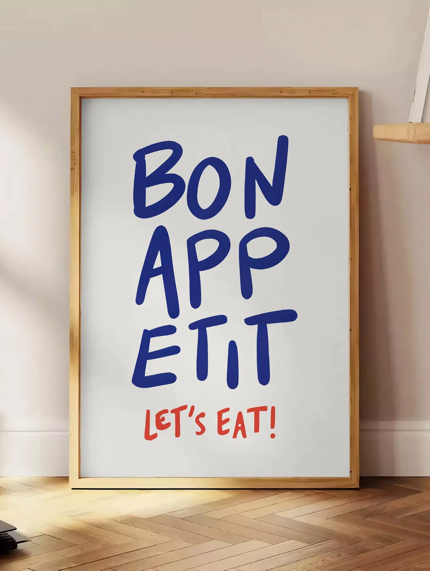 Estampe affiche cuisine Bon Appetit, posee au sol dans un coin salon, sur un parquet a chevrons, pres d'une table d'appoint en chene chargee de livres
