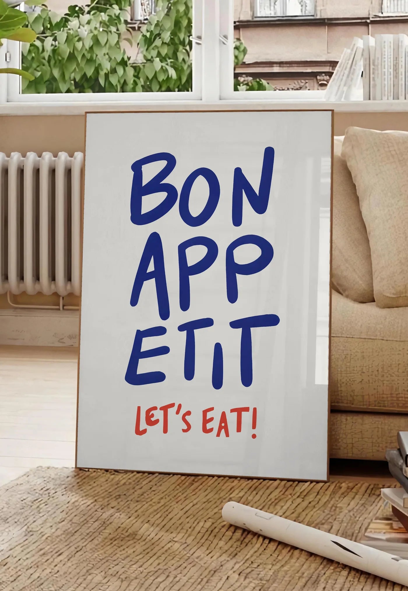 Poster affiche cuisine Bon Appetit, posee au sol dans un salon parisien, devant une grande fenetre donnant sur un immeuble haussmannien, entre un ficus et un canape creme