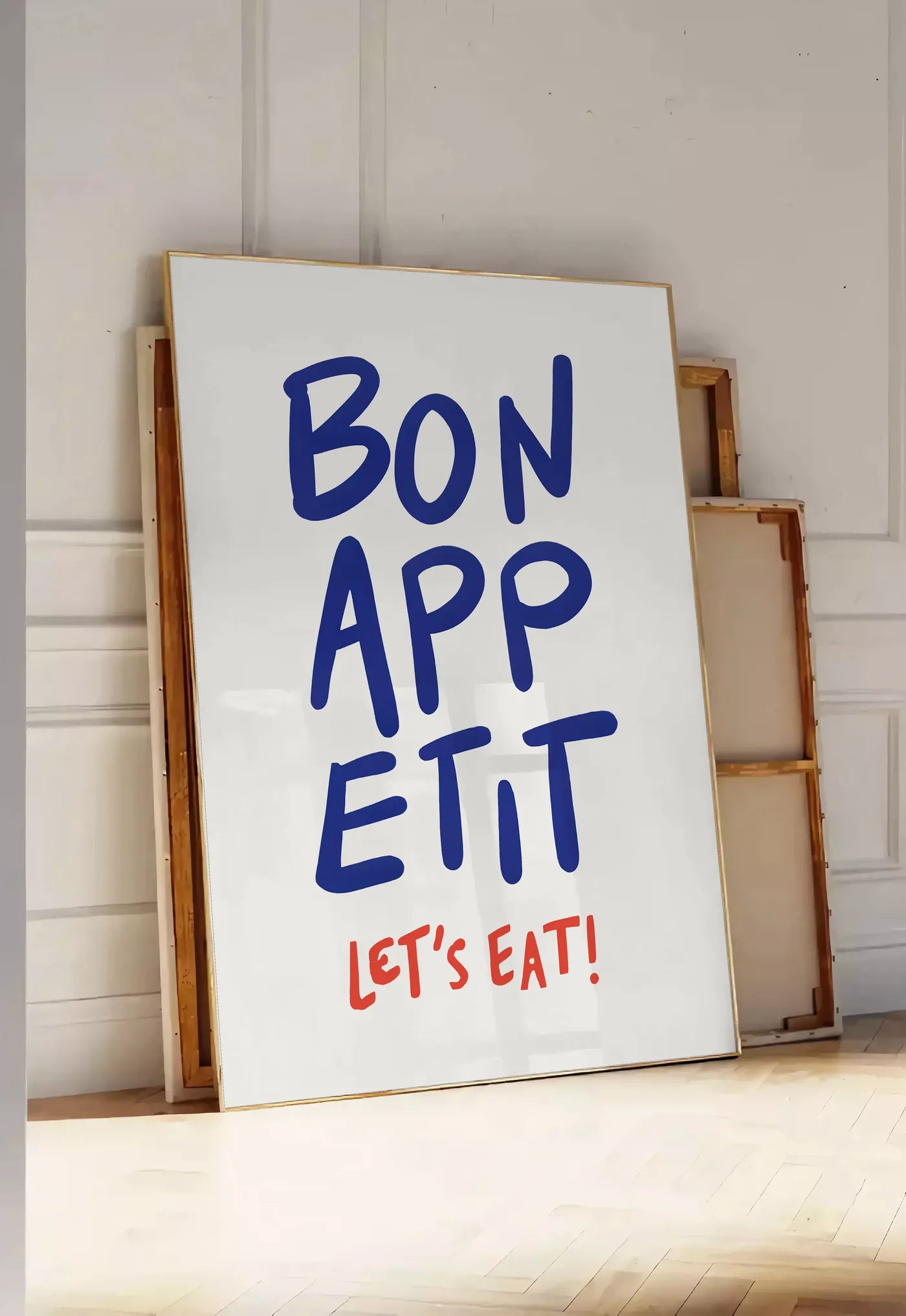 Tirage encadrable affiche cuisine Bon Appetit, posee au sol dans une entree lumineuse, contre un mur a panneaux blancs, a cote de chassis en bois empiles