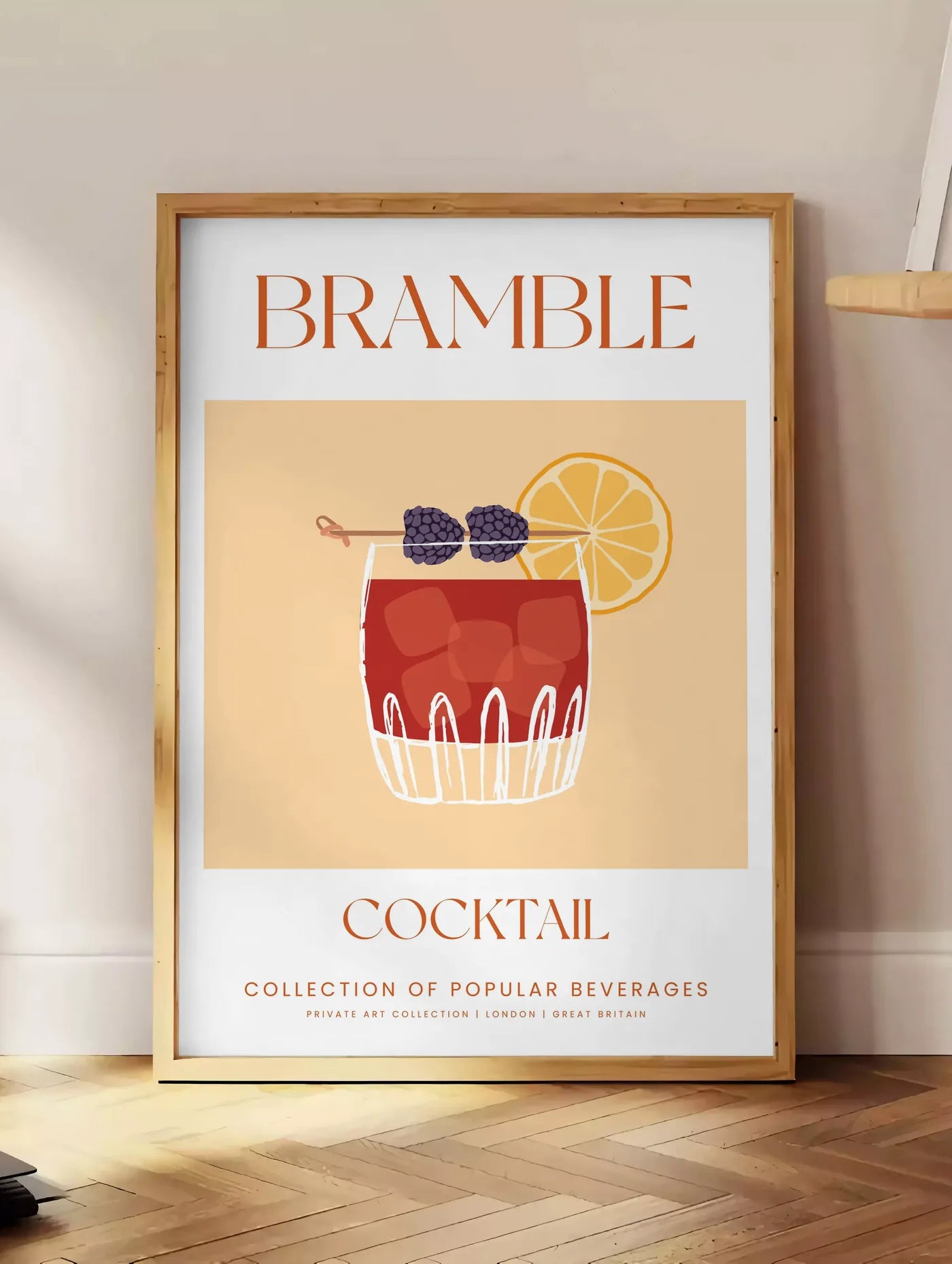 Estampe affiche cocktail Bramble, posee au sol dans un coin salon, sur un parquet a chevrons, pres d'une table d'appoint en chene chargee de livres