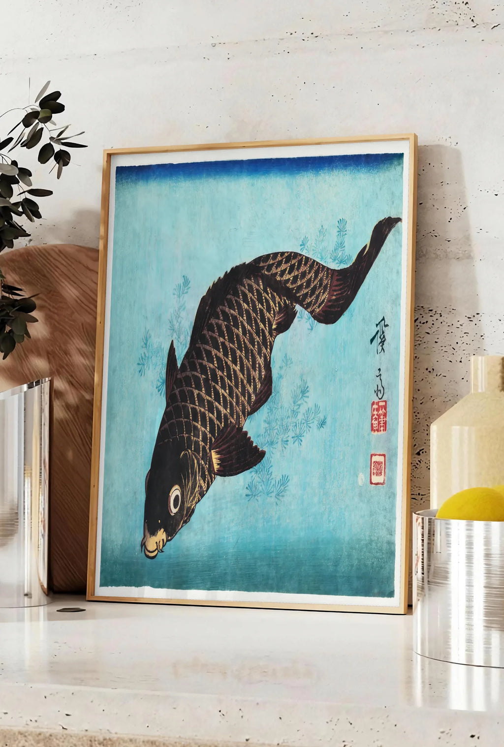 Affiche affiche carpe noire sur fond bleu art japonais, posee sur un plan de travail de cuisine en pierre claire, entre une branche d'eucalyptus en vase et un bocal de citrons