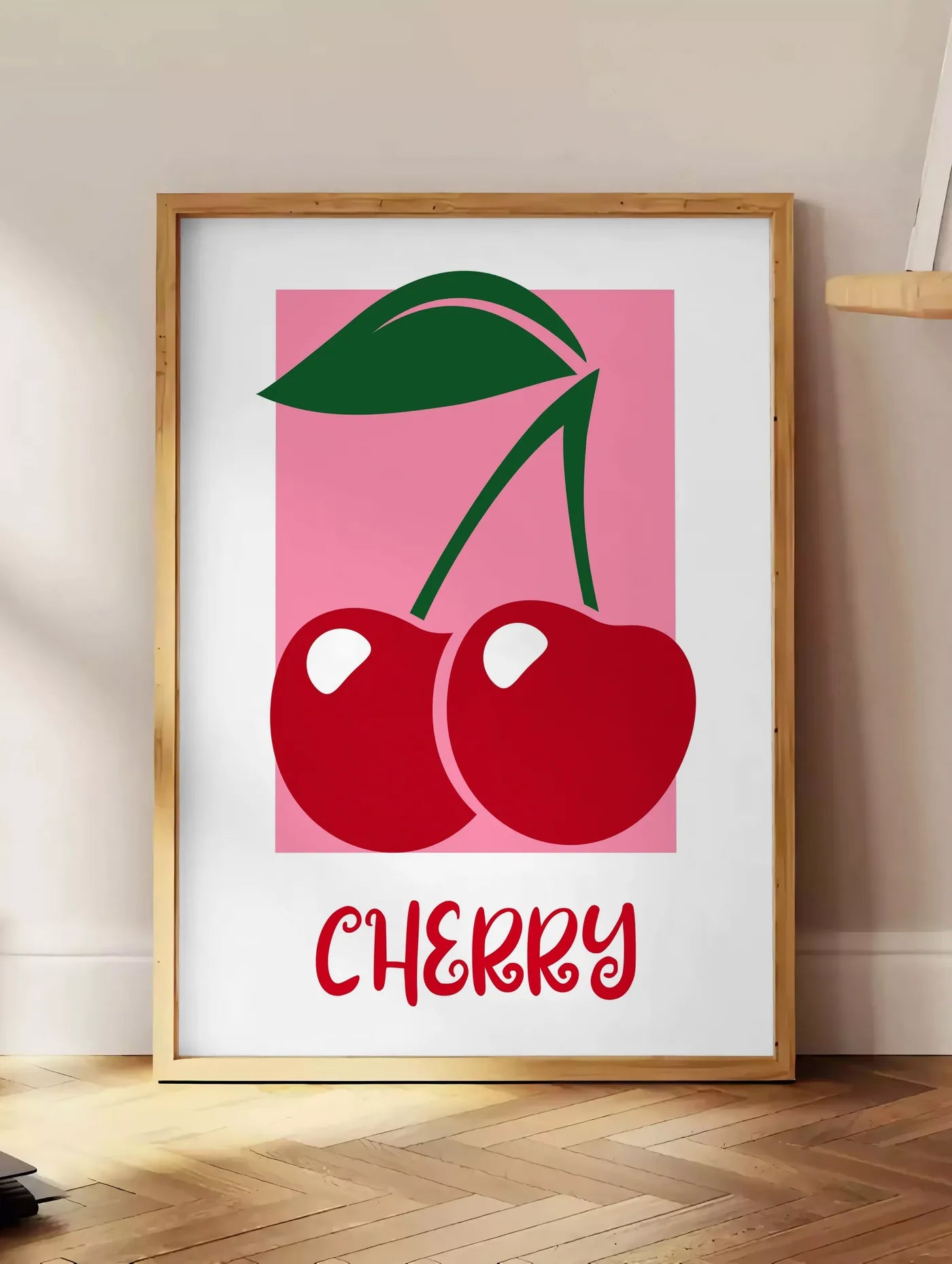 Estampe affiche cuisine cerises cherry fruit rouges et feuille verte sur panneau rose, posee au sol dans un coin salon, sur un parquet a chevrons, pres d'une table d'appoint en chene chargee de livres