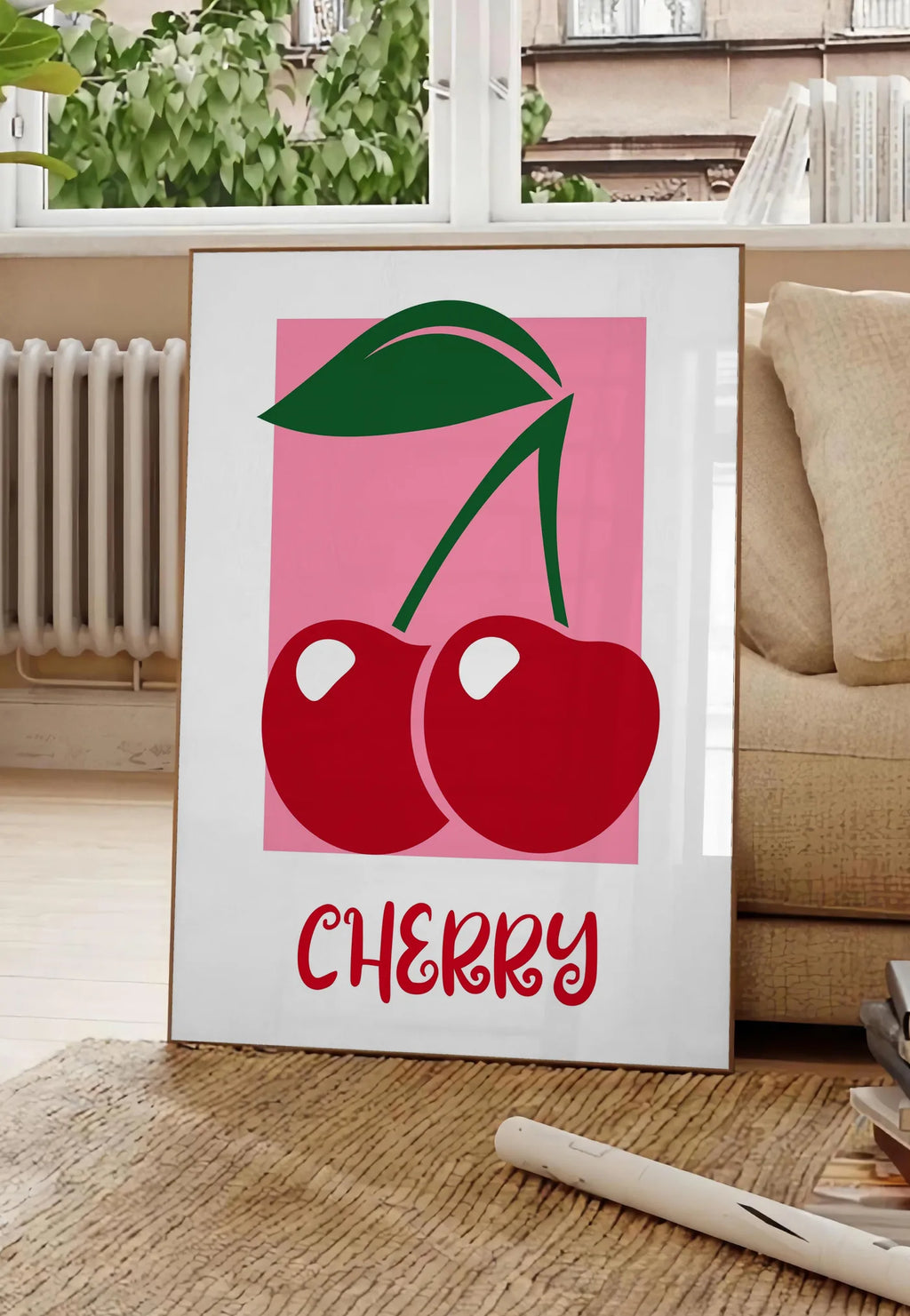 Poster affiche cuisine cerises cherry fruit rouges et feuille verte sur panneau rose, posee au sol dans un salon parisien, devant une grande fenetre donnant sur un immeuble haussmannien, entre un ficus et un canape creme