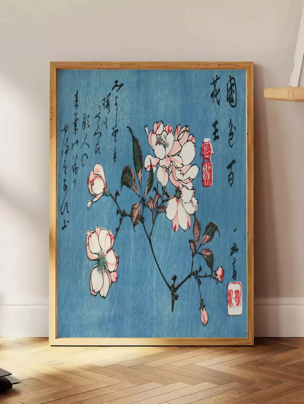 Estampe affiche cerisier en fleurs sur fond bleu art japonais, posee au sol dans un coin salon, sur un parquet a chevrons, pres d'une table d'appoint en chene chargee de livres