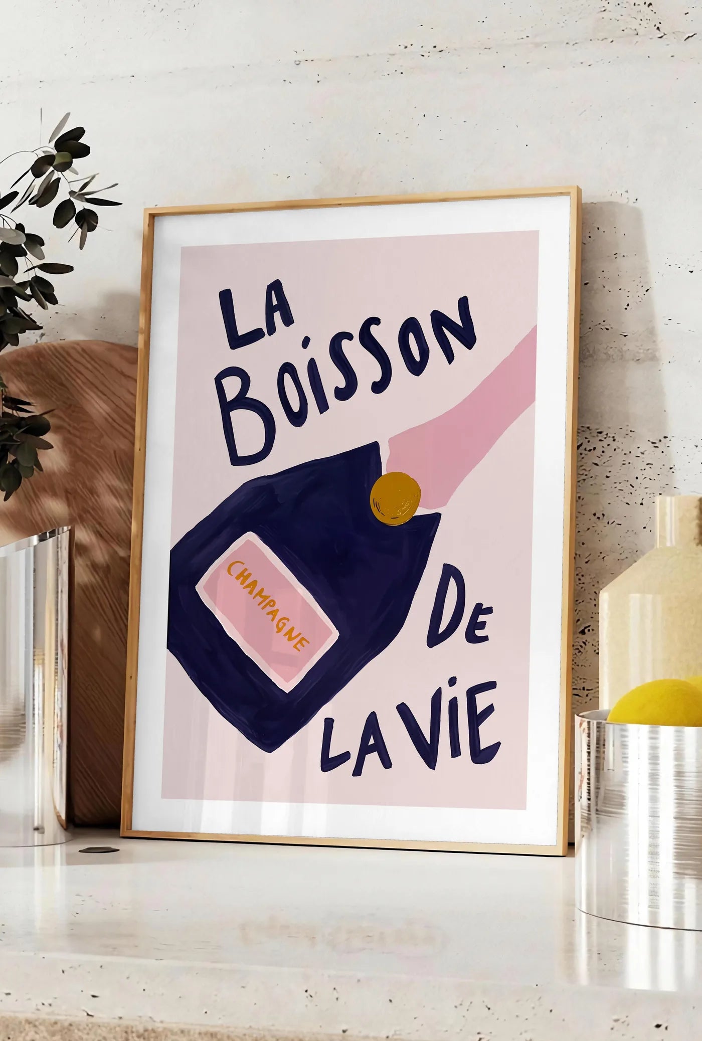 Affiche affiche champagne La Boisson de la Vie, posee sur un plan de travail de cuisine en pierre claire, entre une branche d'eucalyptus en vase et un bocal de citrons