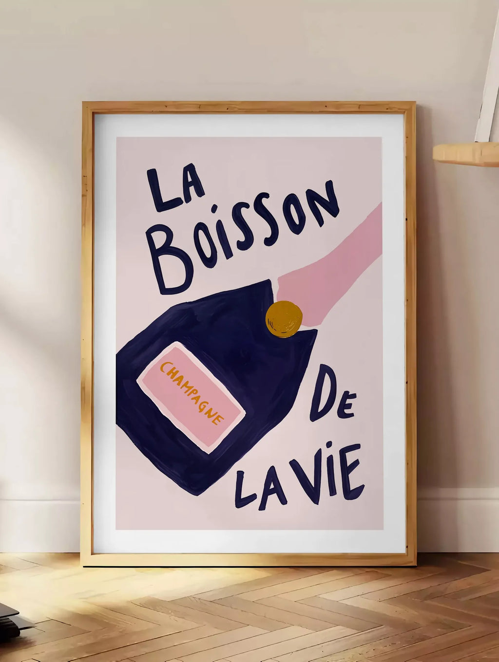 Estampe affiche champagne La Boisson de la Vie, posee au sol dans un coin salon, sur un parquet a chevrons, pres d'une table d'appoint en chene chargee de livres