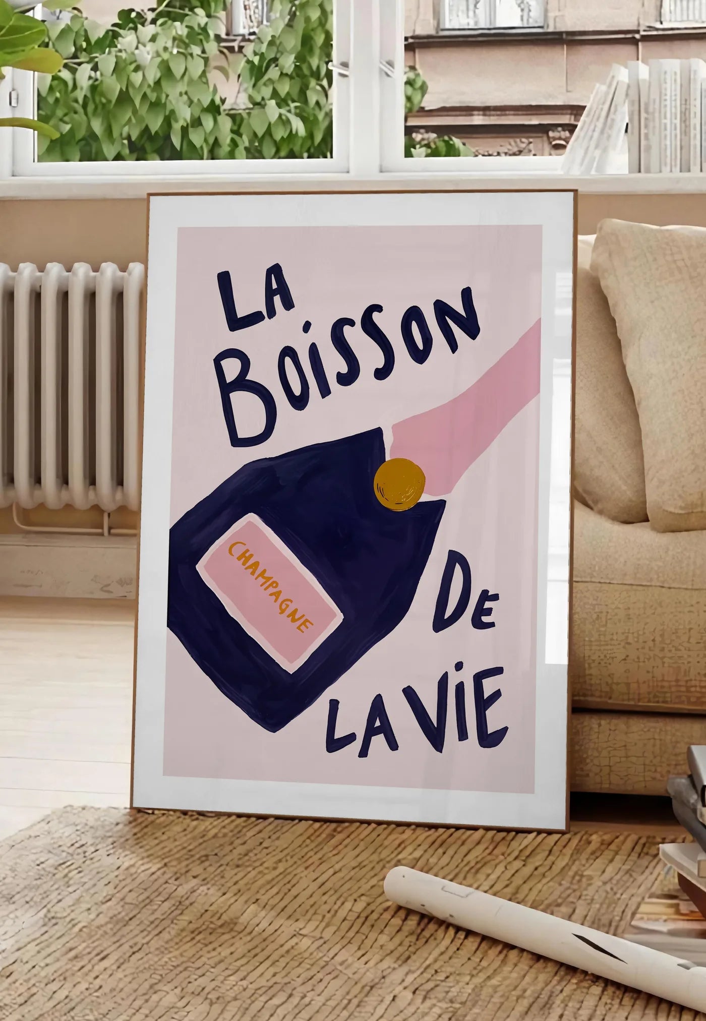 Poster affiche champagne La Boisson de la Vie, posee au sol dans un salon parisien, devant une grande fenetre donnant sur un immeuble haussmannien, entre un ficus et un canape creme