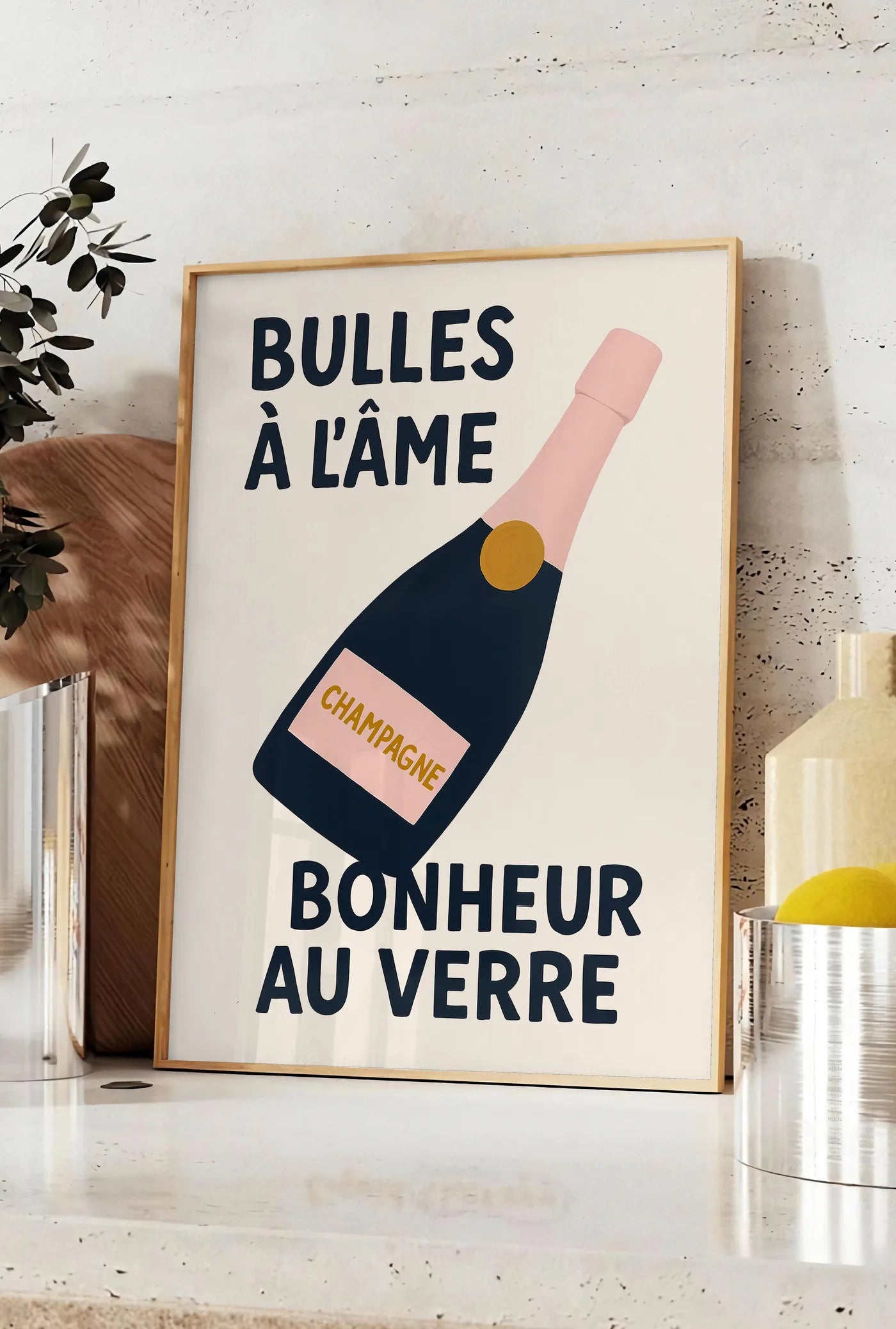 Affiche affiche champagne Bulles a l'ame, posee sur un plan de travail de cuisine en pierre claire, entre une branche d'eucalyptus en vase et un bocal de citrons