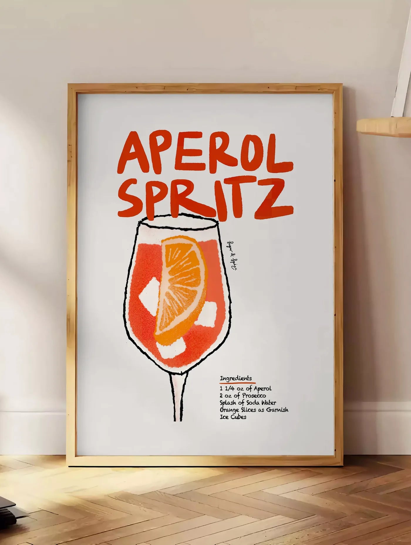 Estampe verre d'Aperol Spritz, posee au sol dans un coin salon, sur un parquet a chevrons, pres d'une table d'appoint en chene chargee de livres