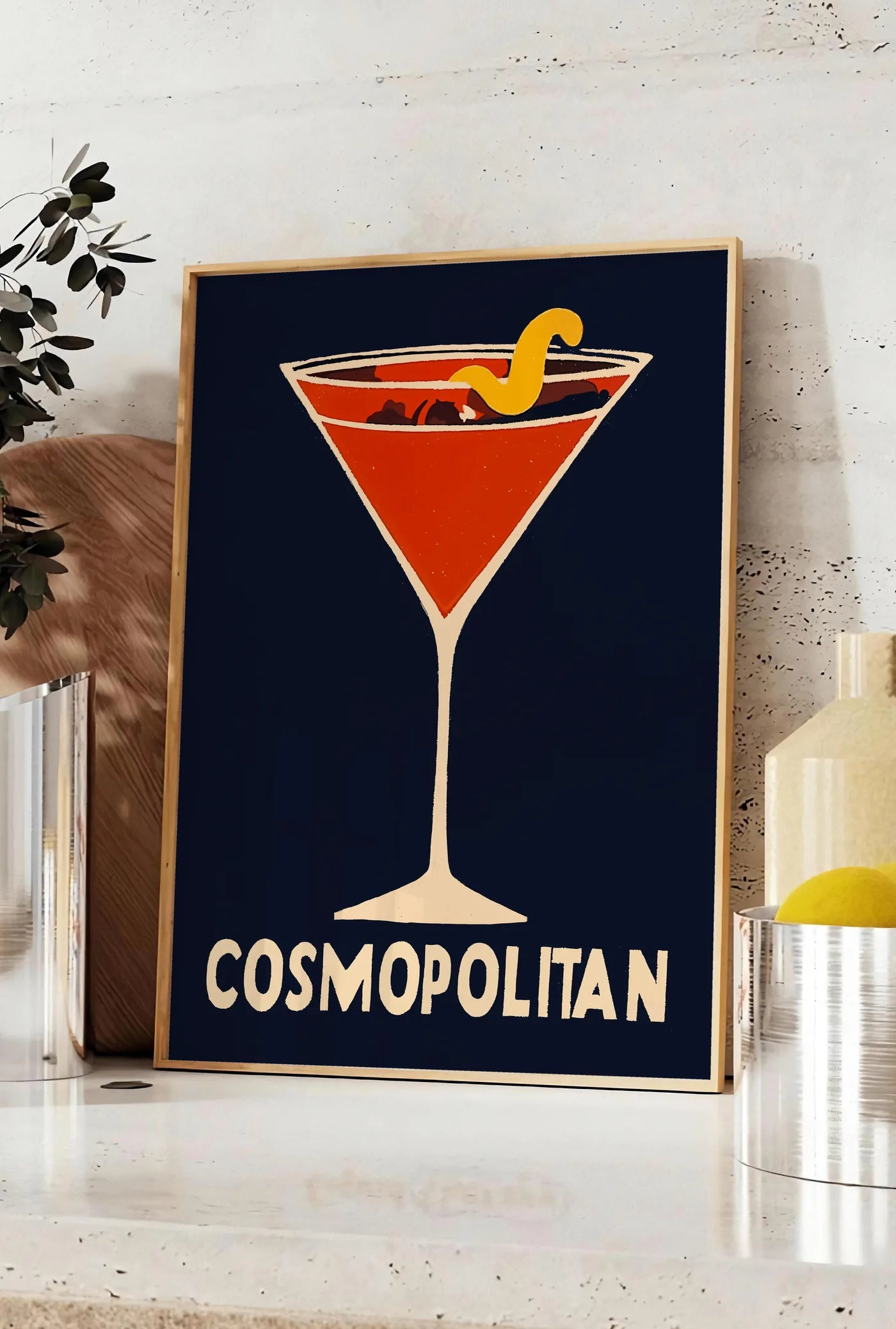 Affiche affiche cocktail Cosmopolitan, posee sur un plan de travail de cuisine en pierre claire, entre une branche d'eucalyptus en vase et un bocal de citrons