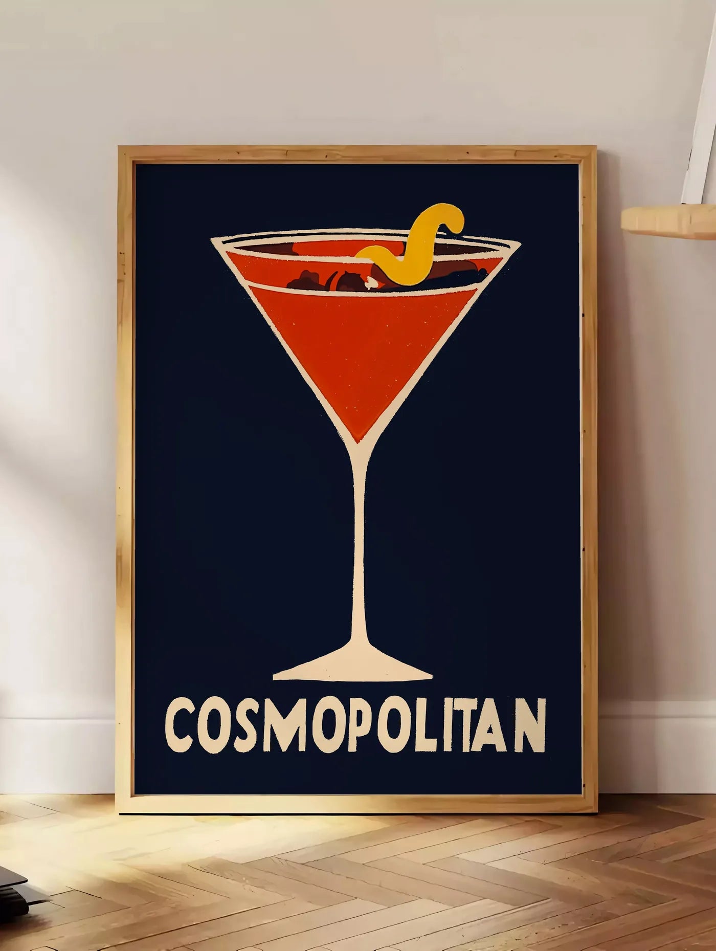Estampe affiche cocktail Cosmopolitan, posee au sol dans un coin salon, sur un parquet a chevrons, pres d'une table d'appoint en chene chargee de livres