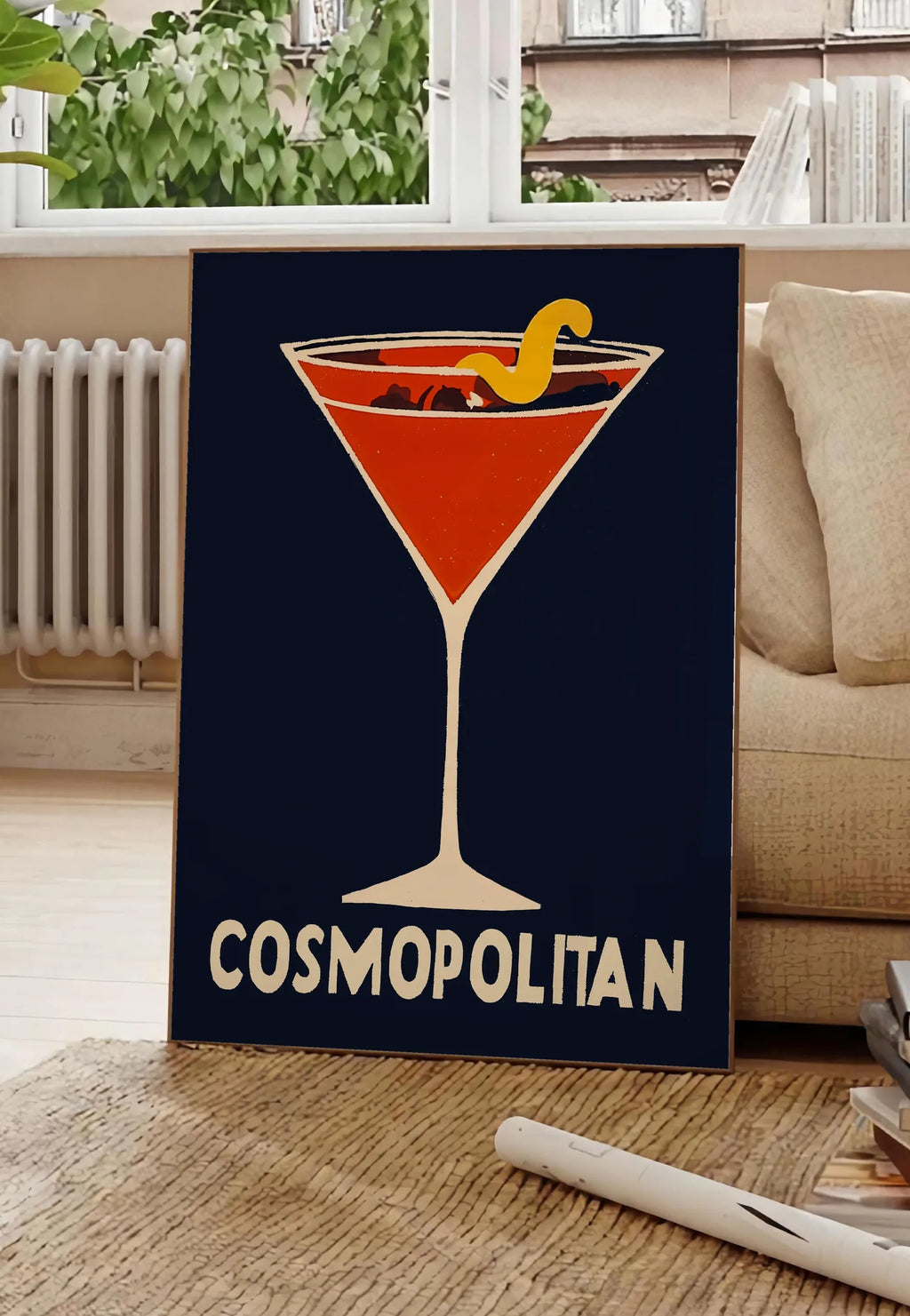 Poster affiche cocktail Cosmopolitan, posee au sol dans un salon parisien, devant une grande fenetre donnant sur un immeuble haussmannien, entre un ficus et un canape creme