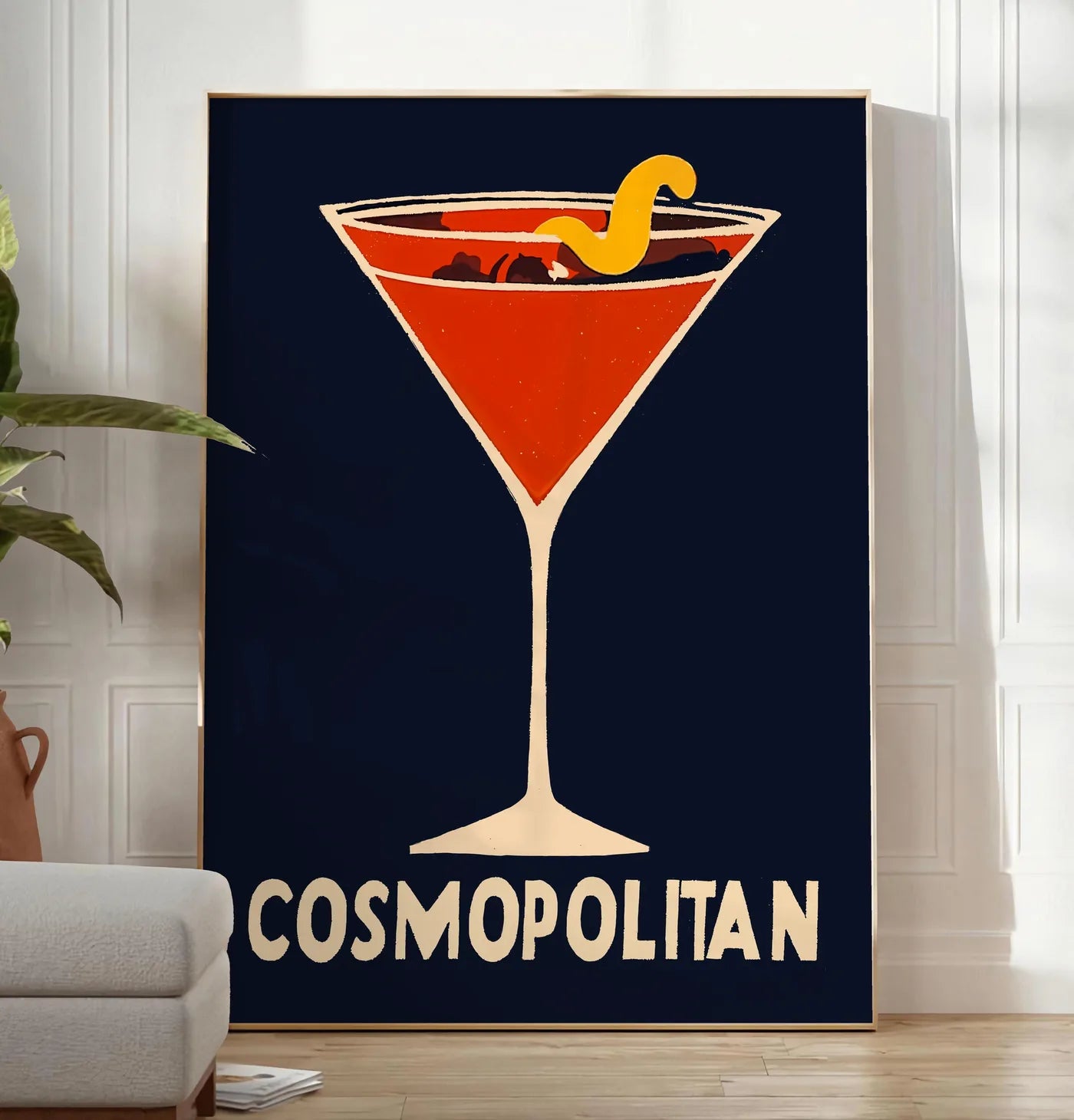 Reproduction affiche cocktail Cosmopolitan, posee au sol dans un salon minimaliste clair, a cote d'un canape beige et d'une plante verte, sur un parquet clair