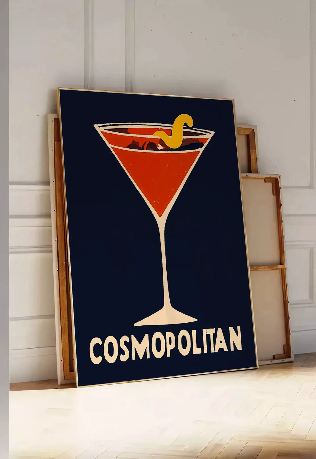 Tirage encadrable affiche cocktail Cosmopolitan, posee au sol dans une entree lumineuse, contre un mur a panneaux blancs, a cote de chassis en bois empiles