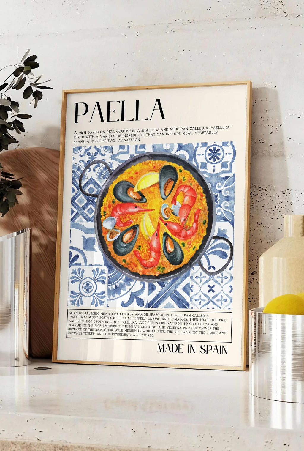 Affiche affiche paella aquarelle, posee sur un plan de travail de cuisine en pierre claire, entre une branche d'eucalyptus en vase et un bocal de citrons