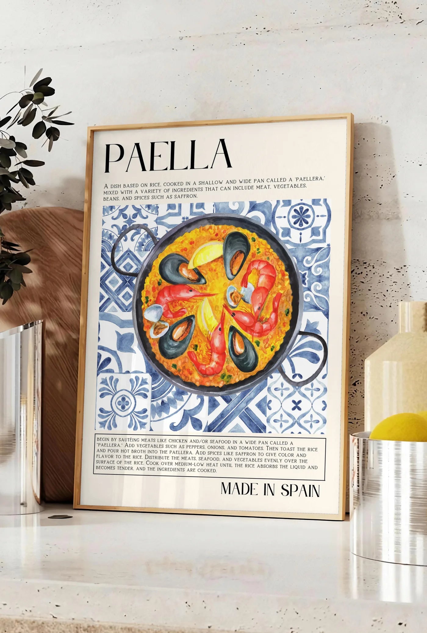 Affiche affiche paella aquarelle, posee sur un plan de travail de cuisine en pierre claire, entre une branche d'eucalyptus en vase et un bocal de citrons