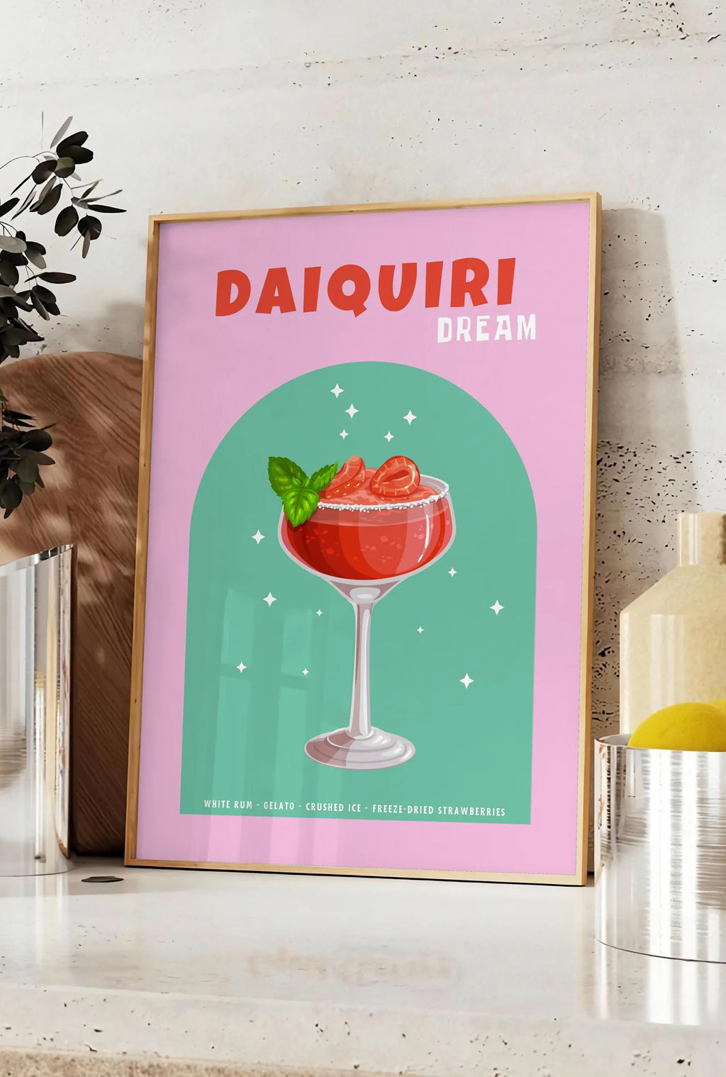 Affiche affiche cocktail Daiquiri fraise, posee sur un plan de travail de cuisine en pierre claire, entre une branche d'eucalyptus en vase et un bocal de citrons