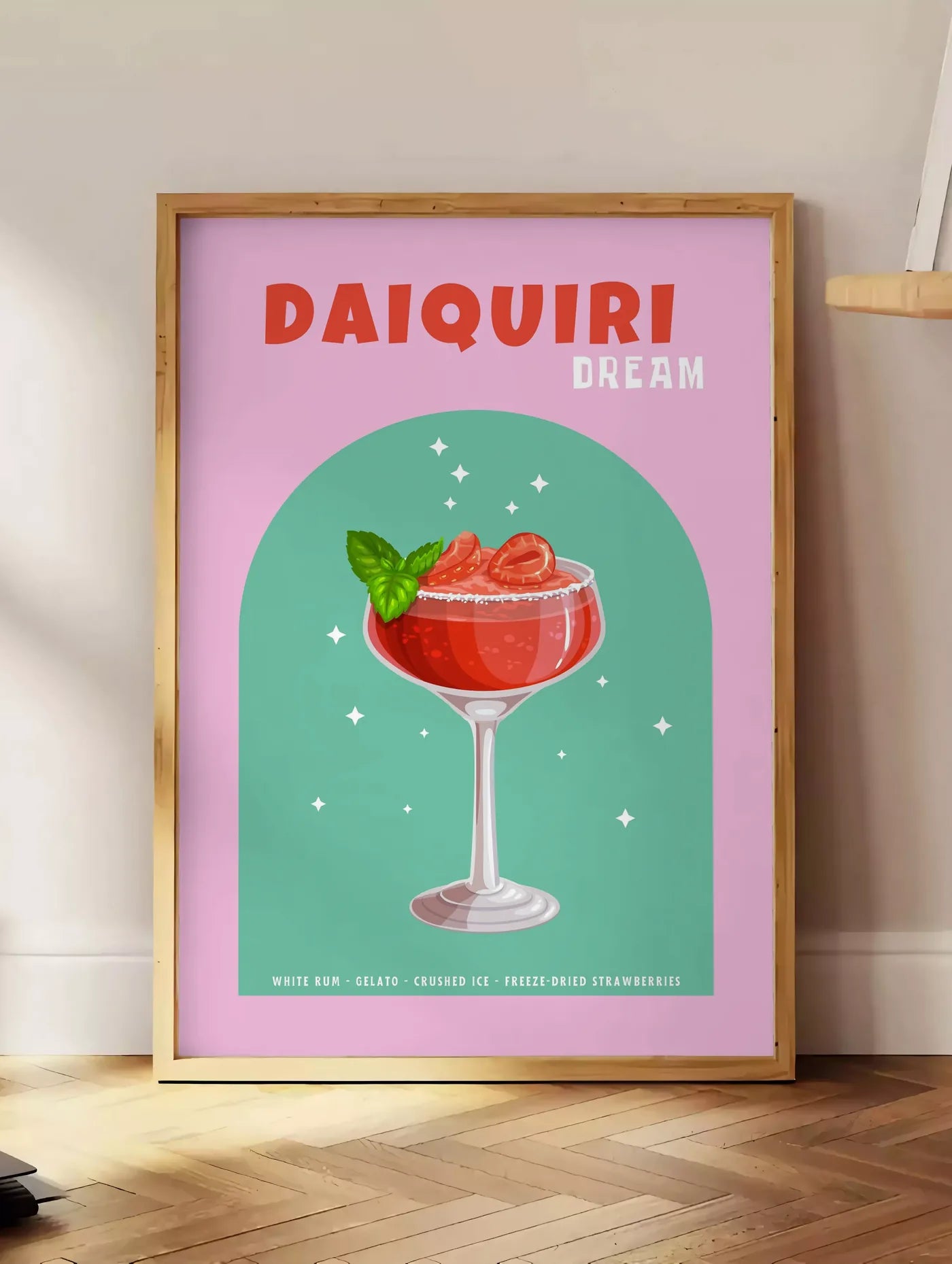 Estampe affiche cocktail Daiquiri fraise, posee au sol dans un coin salon, sur un parquet a chevrons, pres d'une table d'appoint en chene chargee de livres