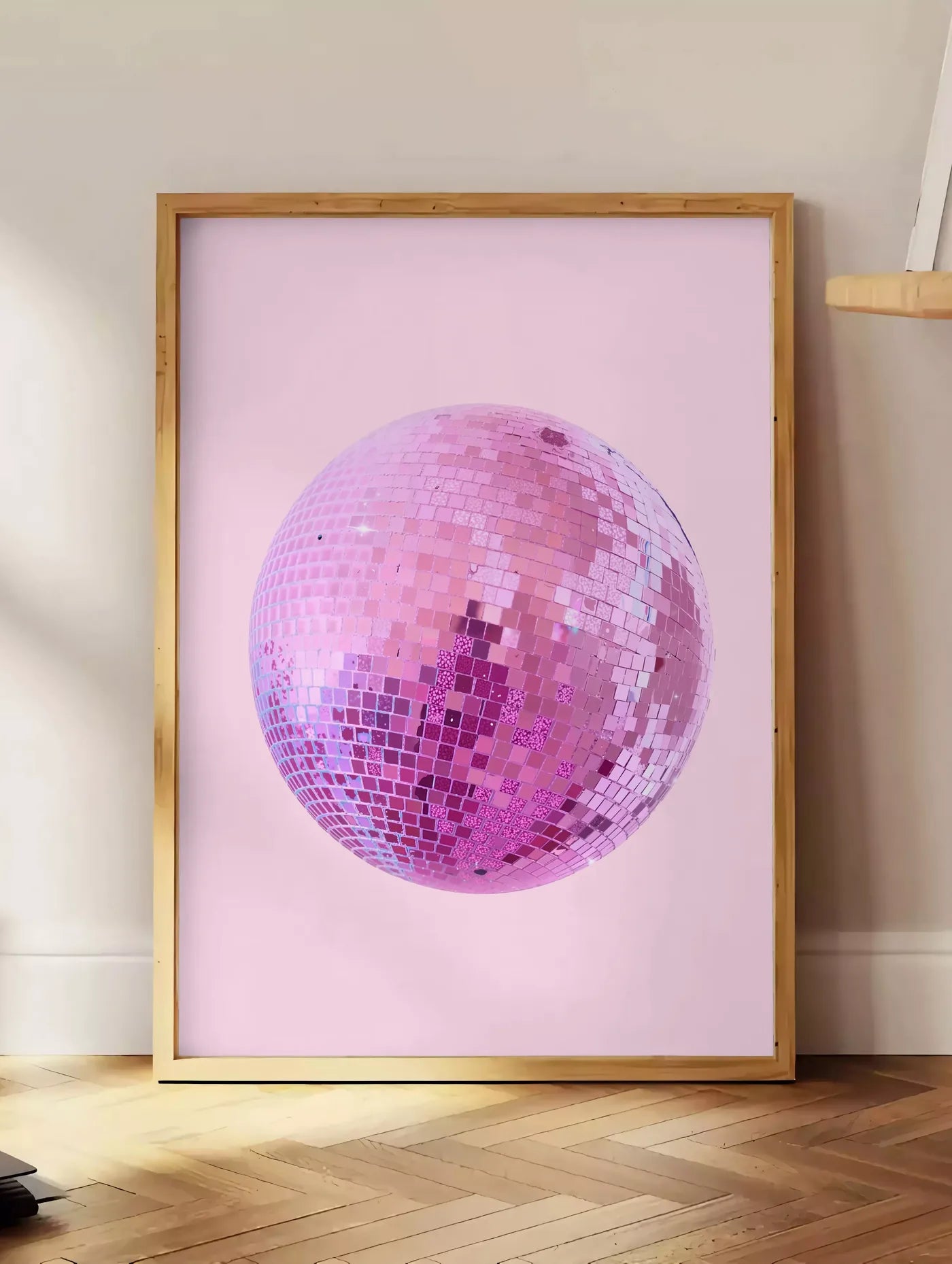 Estampe affiche vintage boule disco rose a facettes sur fond rose poudre, posee au sol dans un coin salon, sur un parquet a chevrons, pres d'une table d'appoint en chene chargee de livres