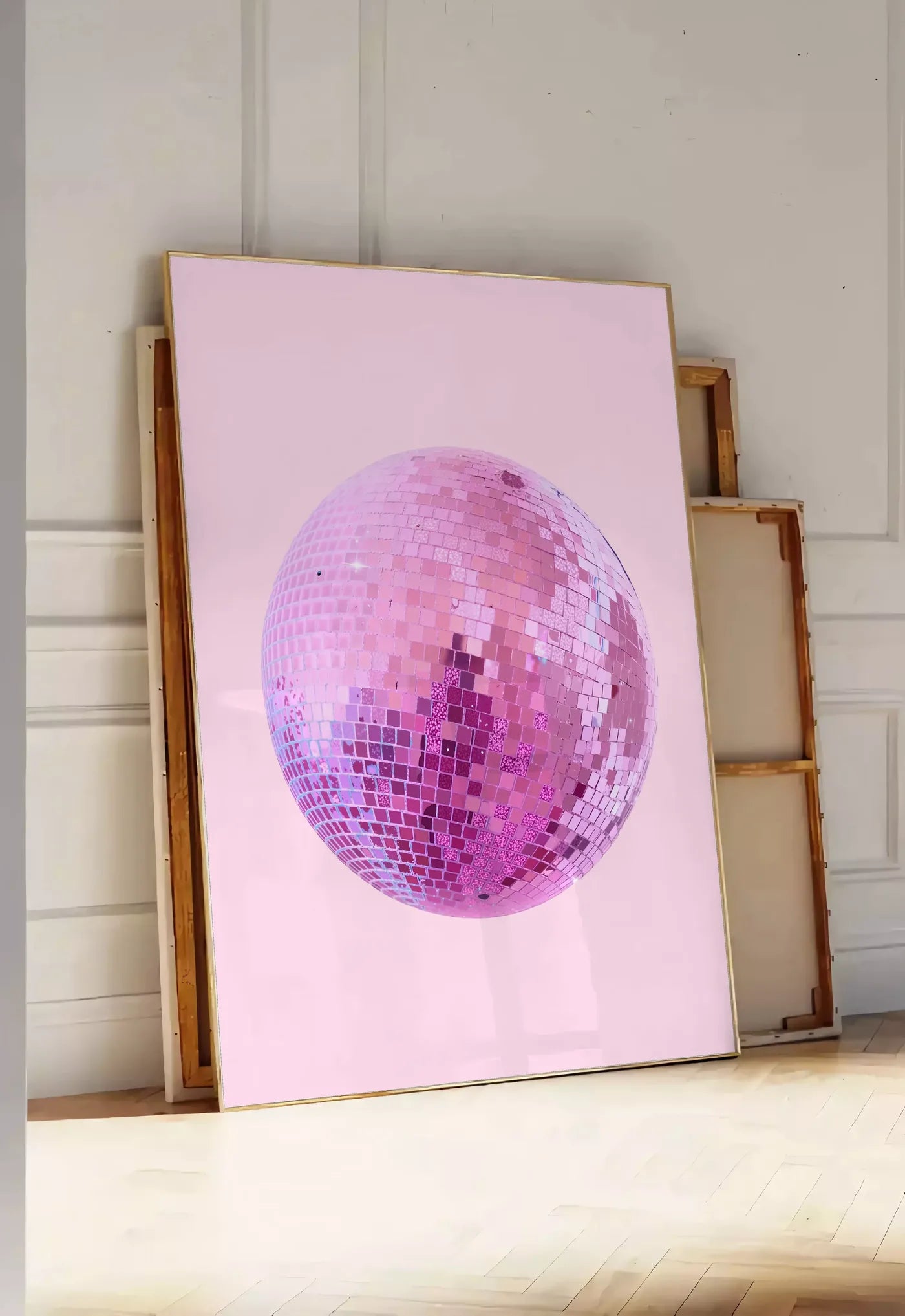 Tirage encadrable affiche vintage boule disco rose a facettes sur fond rose poudre, posee au sol dans une entree lumineuse, contre un mur a panneaux blancs, a cote de chassis en bois empiles