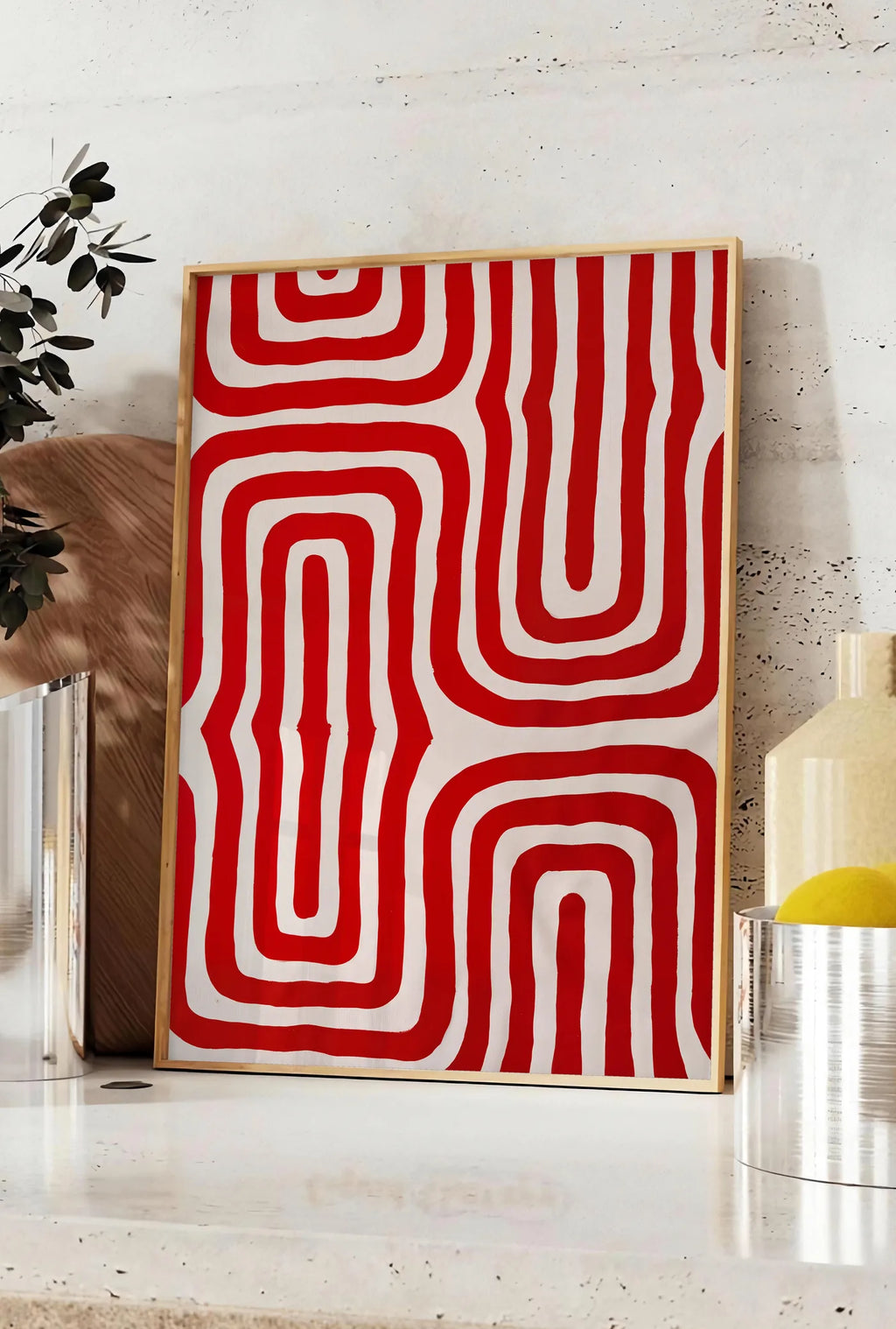 Affiche labyrinthe de lignes rouges ondulees sur fond creme, posee sur un plan de travail de cuisine en pierre claire, entre une branche d'eucalyptus en vase et un bocal de citrons
