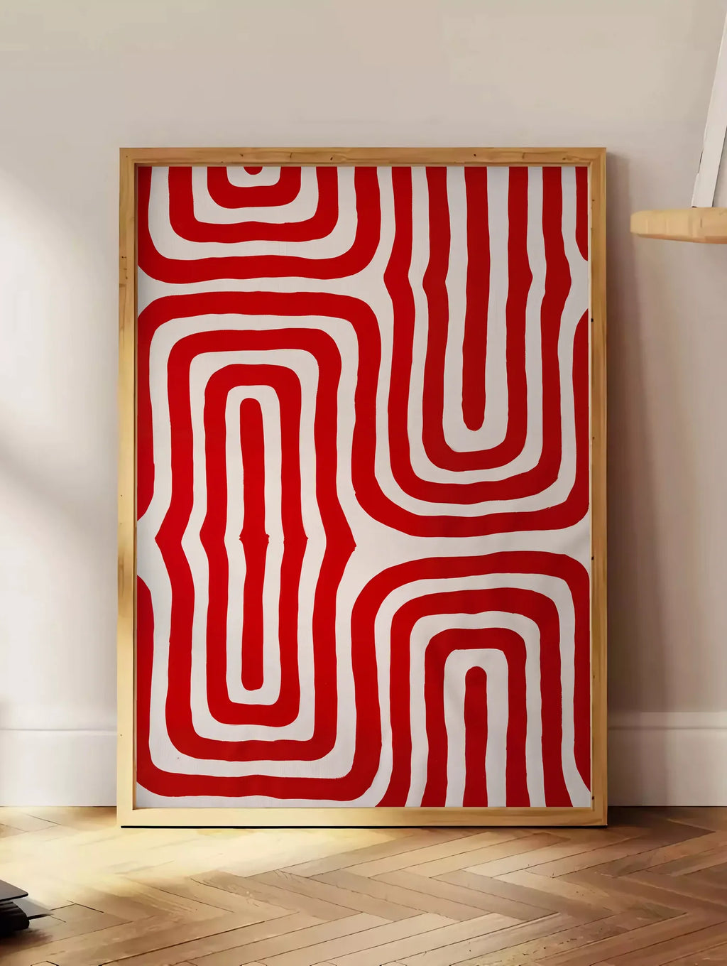 Estampe labyrinthe de lignes rouges ondulees sur fond creme, posee au sol dans un coin salon, sur un parquet a chevrons, pres d'une table d'appoint en chene chargee de livres