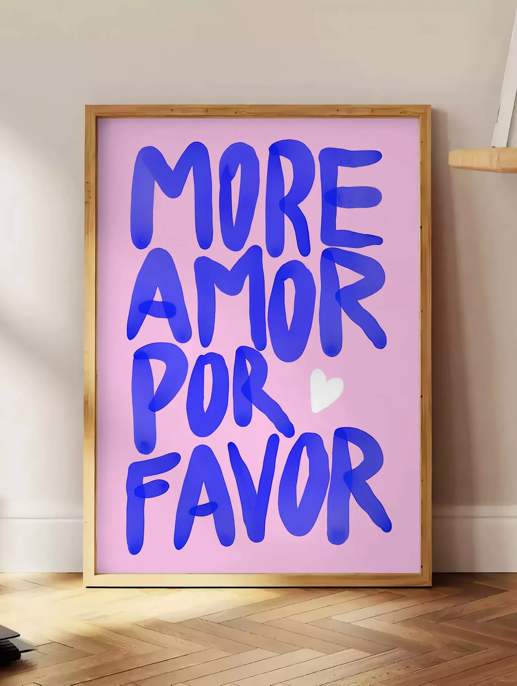 Estampe affiche vintage More Amor Por Favor, posee au sol dans un coin salon, sur un parquet a chevrons, pres d'une table d'appoint en chene chargee de livres