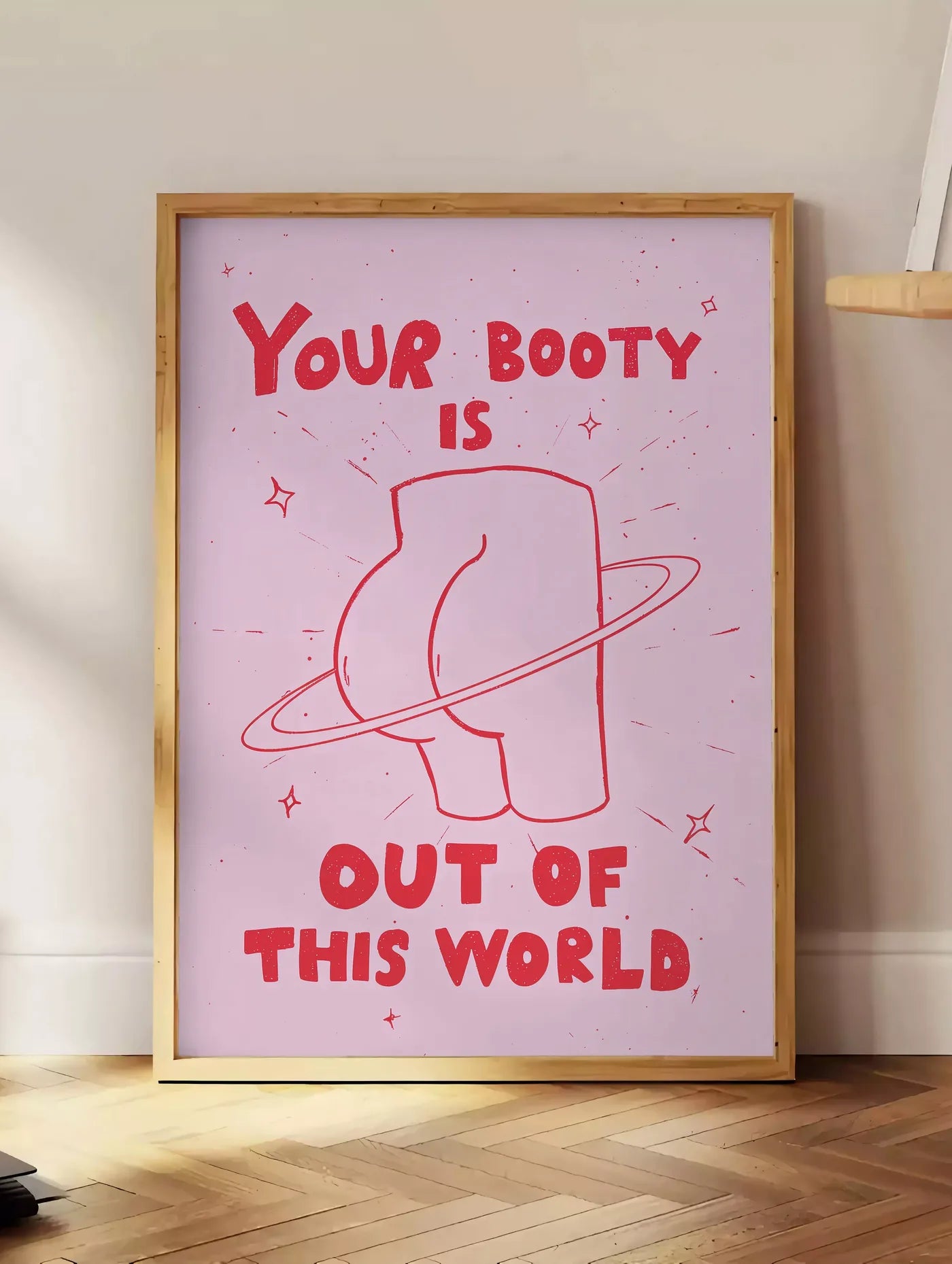 Estampe affiche vintage Your Booty, posee au sol dans un coin salon, sur un parquet a chevrons, pres d'une table d'appoint en chene chargee de livres