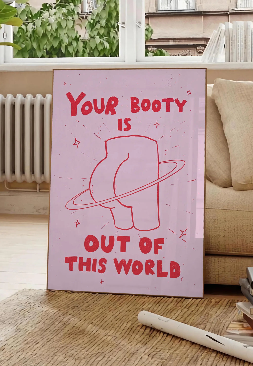Poster affiche vintage Your Booty, posee au sol dans un salon parisien, devant une grande fenetre donnant sur un immeuble haussmannien, entre un ficus et un canape creme