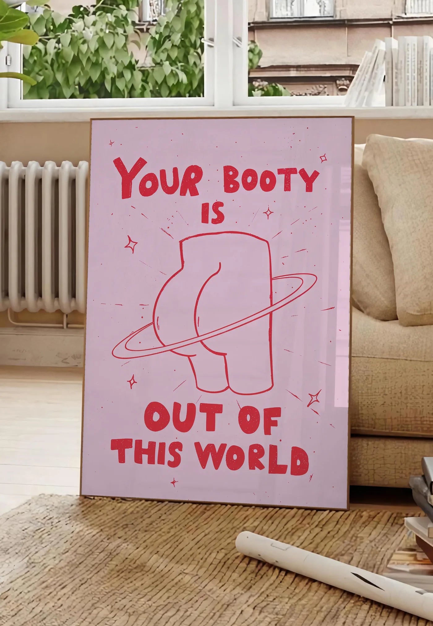 Poster affiche vintage Your Booty, posee au sol dans un salon parisien, devant une grande fenetre donnant sur un immeuble haussmannien, entre un ficus et un canape creme