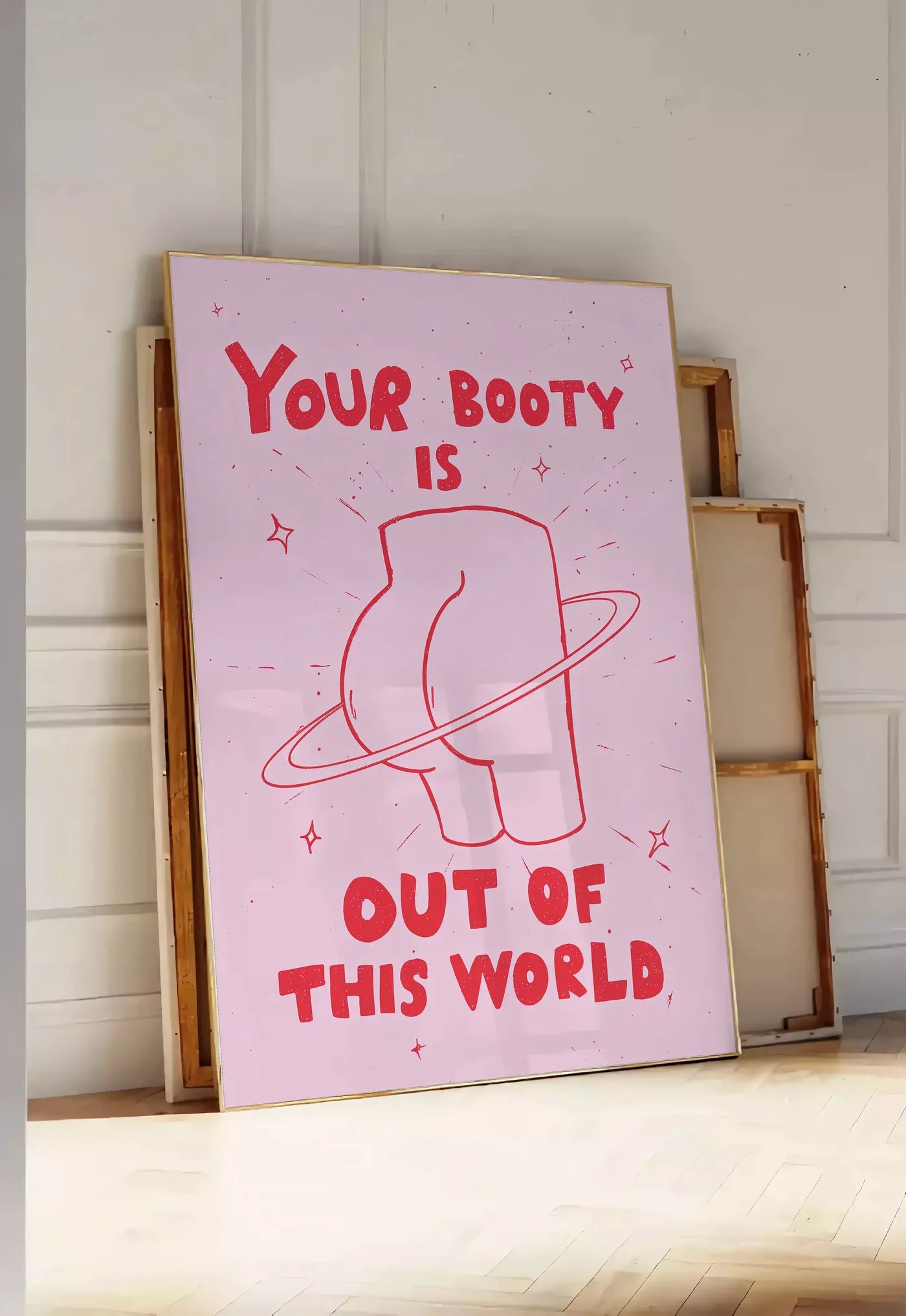 Tirage encadrable affiche vintage Your Booty, posee au sol dans une entree lumineuse, contre un mur a panneaux blancs, a cote de chassis en bois empiles
