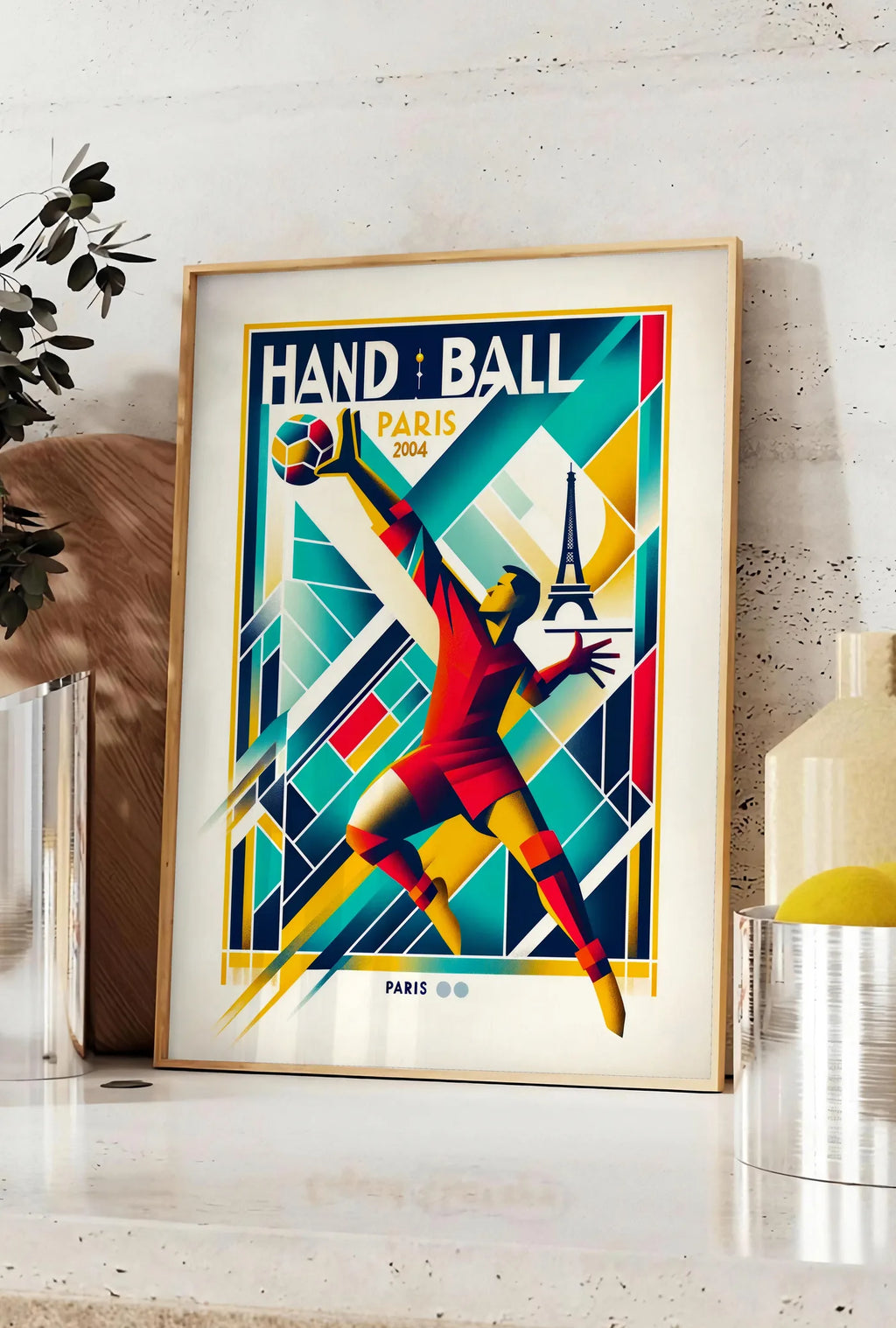 Affiche stylisé joueur de handball en saut, posee sur un plan de travail de cuisine en pierre claire, entre une branche d'eucalyptus en vase et un bocal de citrons