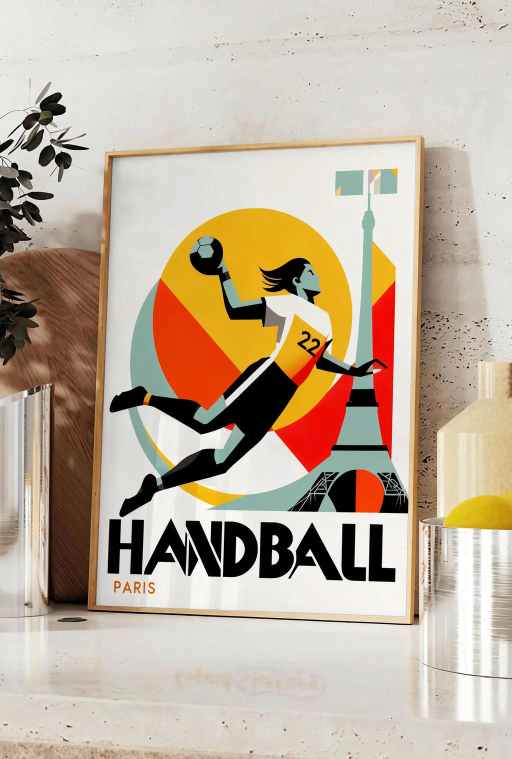 Affiche affiche handball Paris, posee sur un plan de travail de cuisine en pierre claire, entre une branche d'eucalyptus en vase et un bocal de citrons